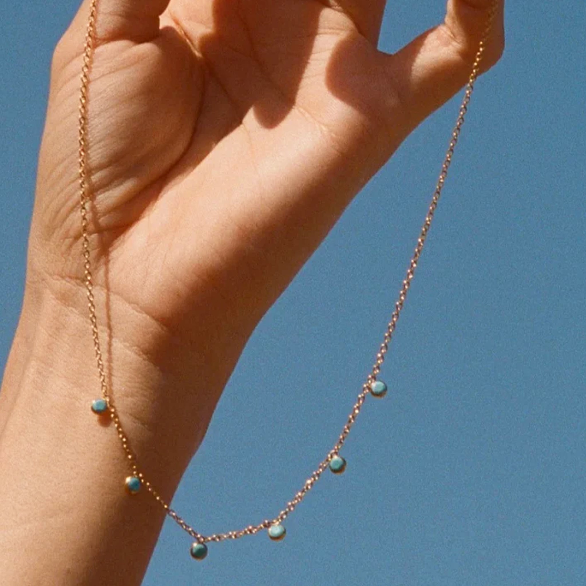 Hand holding a delicate necklace with blue beads against a clear blue sky
