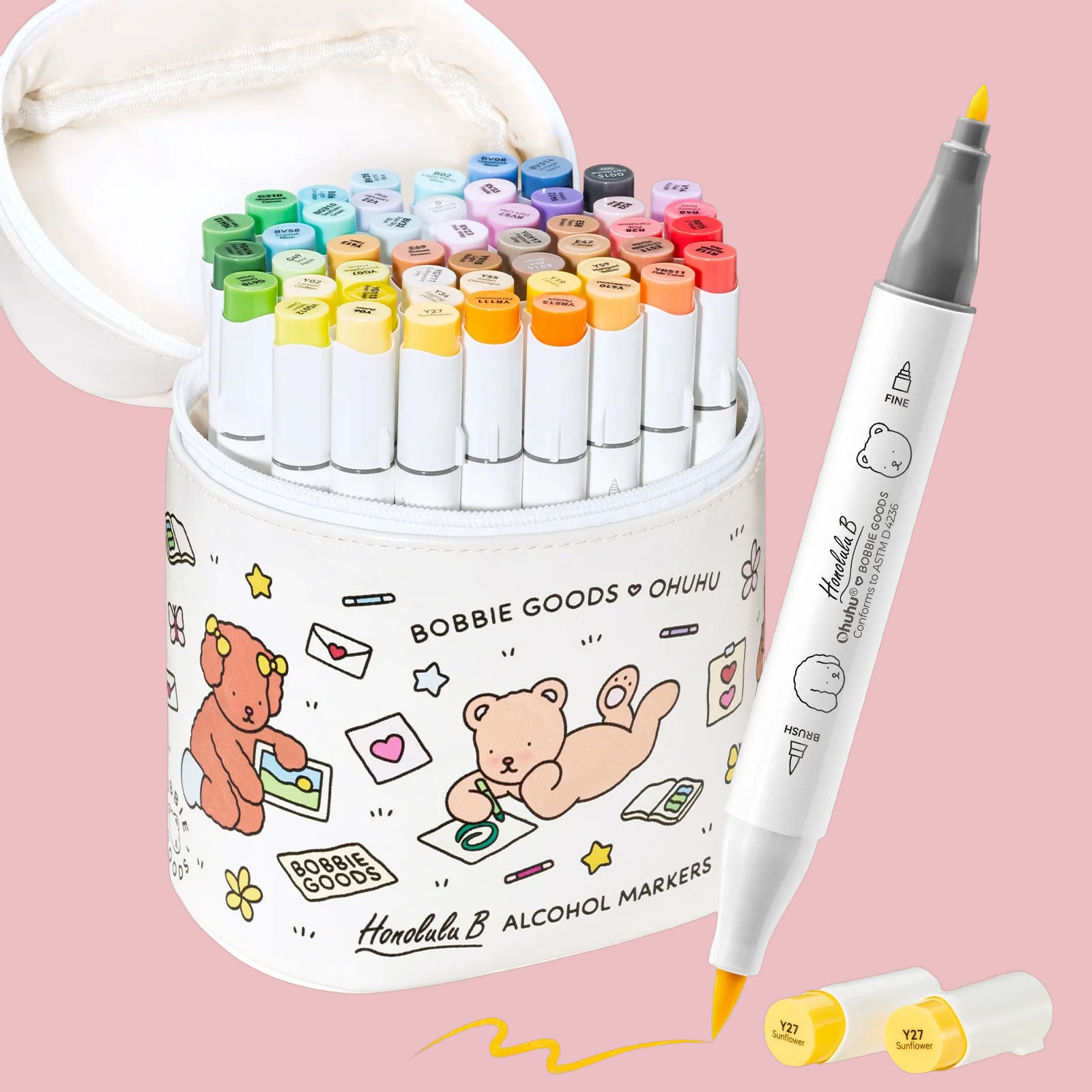 An open white Bobbie Goods x Ohuhu marker pouch packed with dozens of markers arranged by color, with a single dual-tip marker and two yellow caps placed beside it on a pink backdrop.