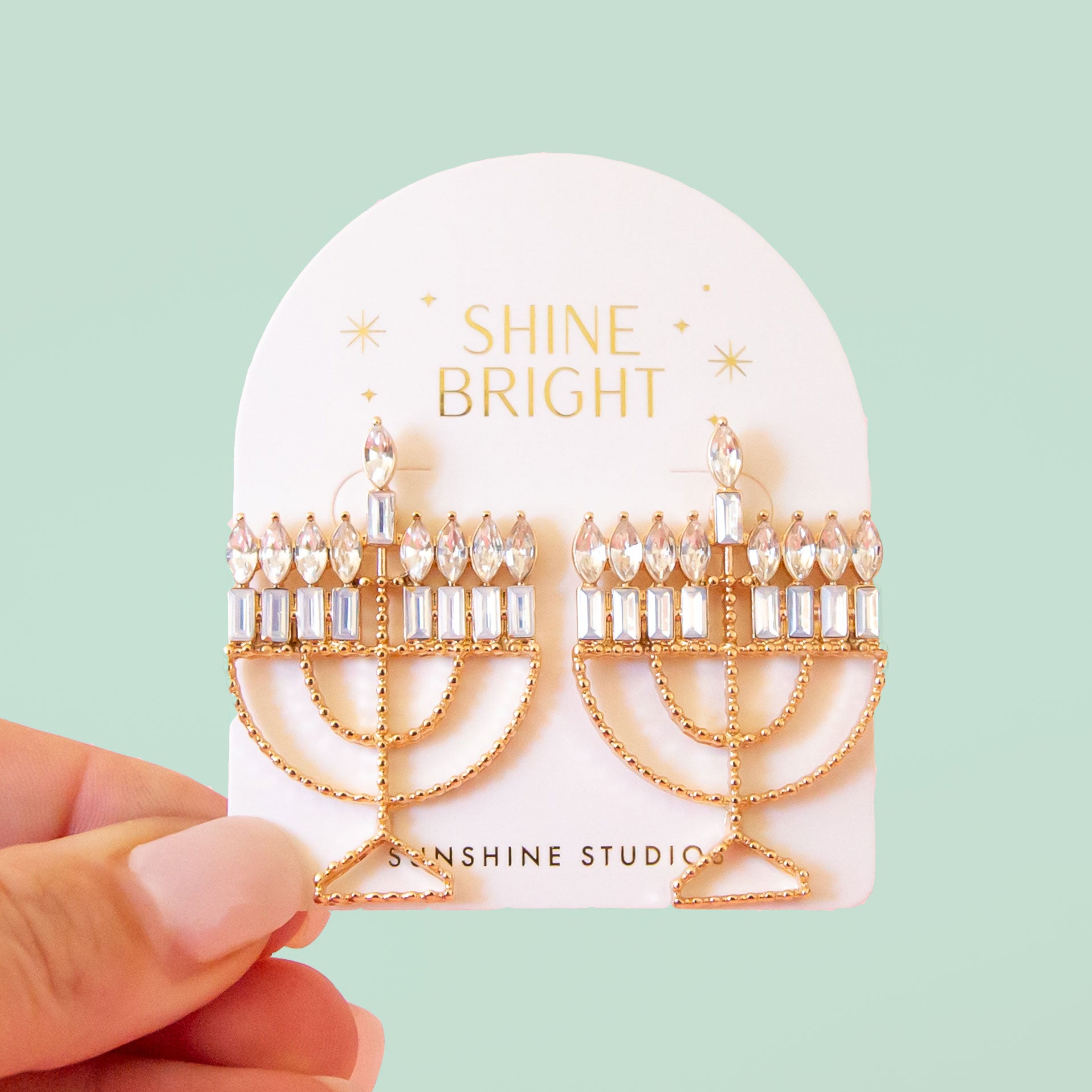 Menorah shaped gold earrings are attached to a cream colored card. the Card has gold foil stamp which reads Shine Bright and Sunshine Studios with glittering stars surrounding the text. The background of the photo is a soft warm pink color. 