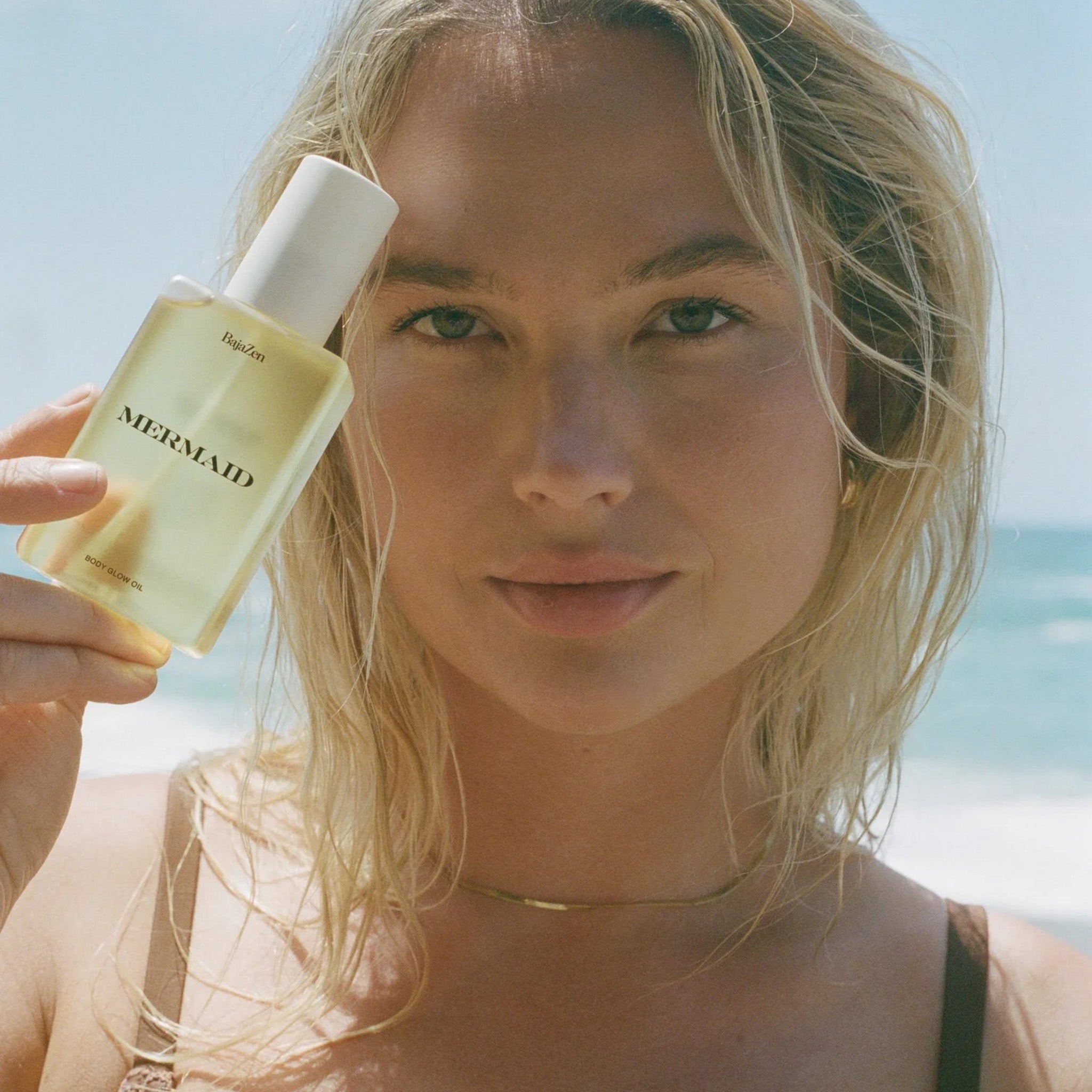 Woman holding MERMAID body glow oil by BajaZen on a beach with the ocean in the background.