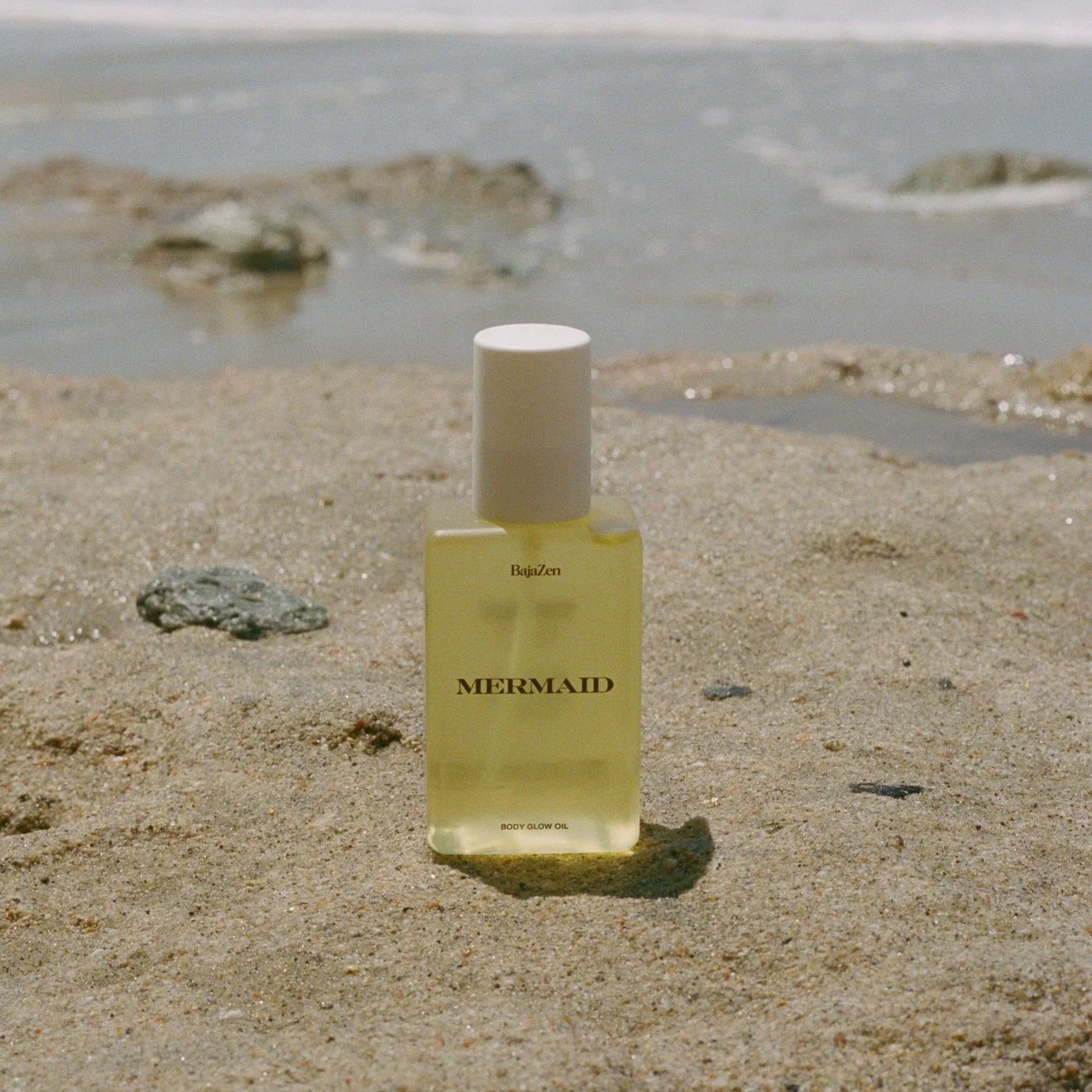 Bottle of MERMAID body glow oil by BajaZen placed on the beach with ocean water in the background.