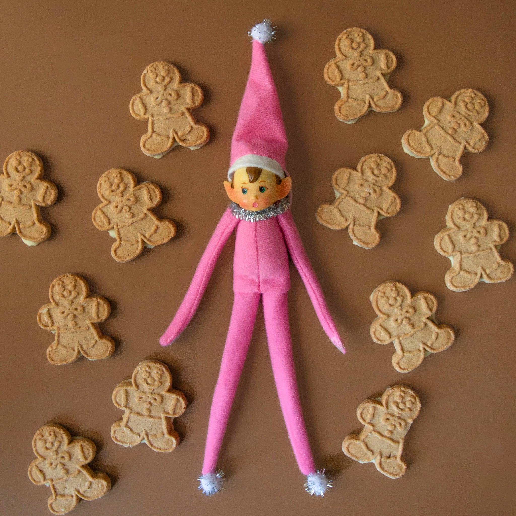 A pink elf ornament lies on a brown background with gingerbread cookies surrounding it.