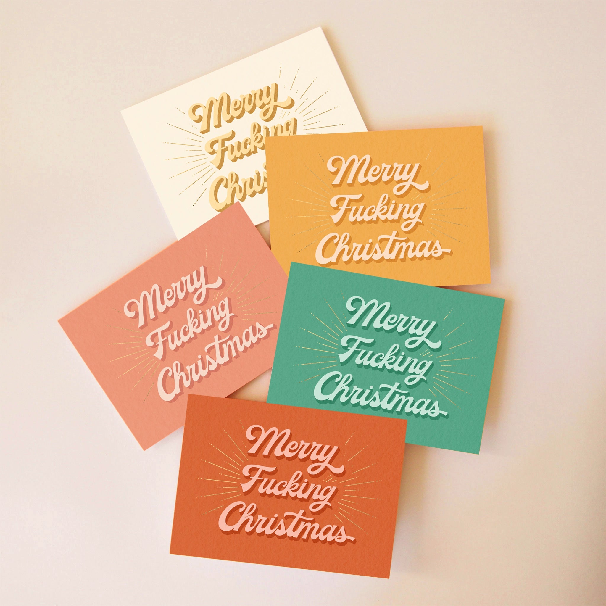 On a beige background is a pack of five holiday greeting cards in a variety of colors including, green, rust, salmon, orange and ivory. The front of each card reads, "Merry Fucking Christmas".