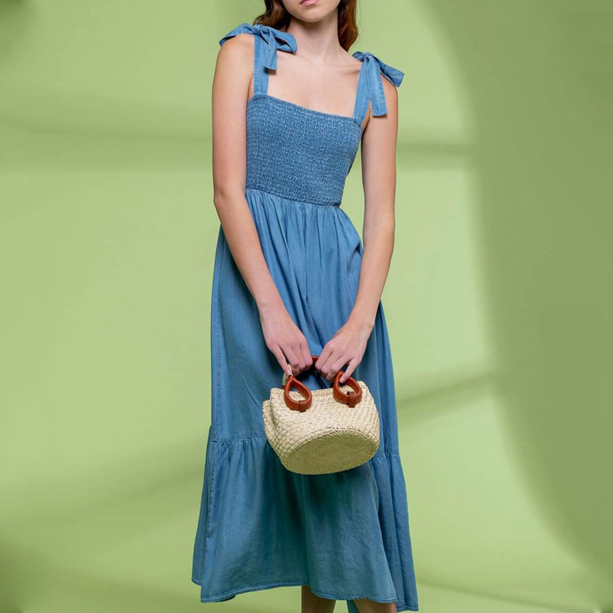 On a green shadow background is a model wearing a blue flowy midi dress with tie shoulder straps and a square neckline.