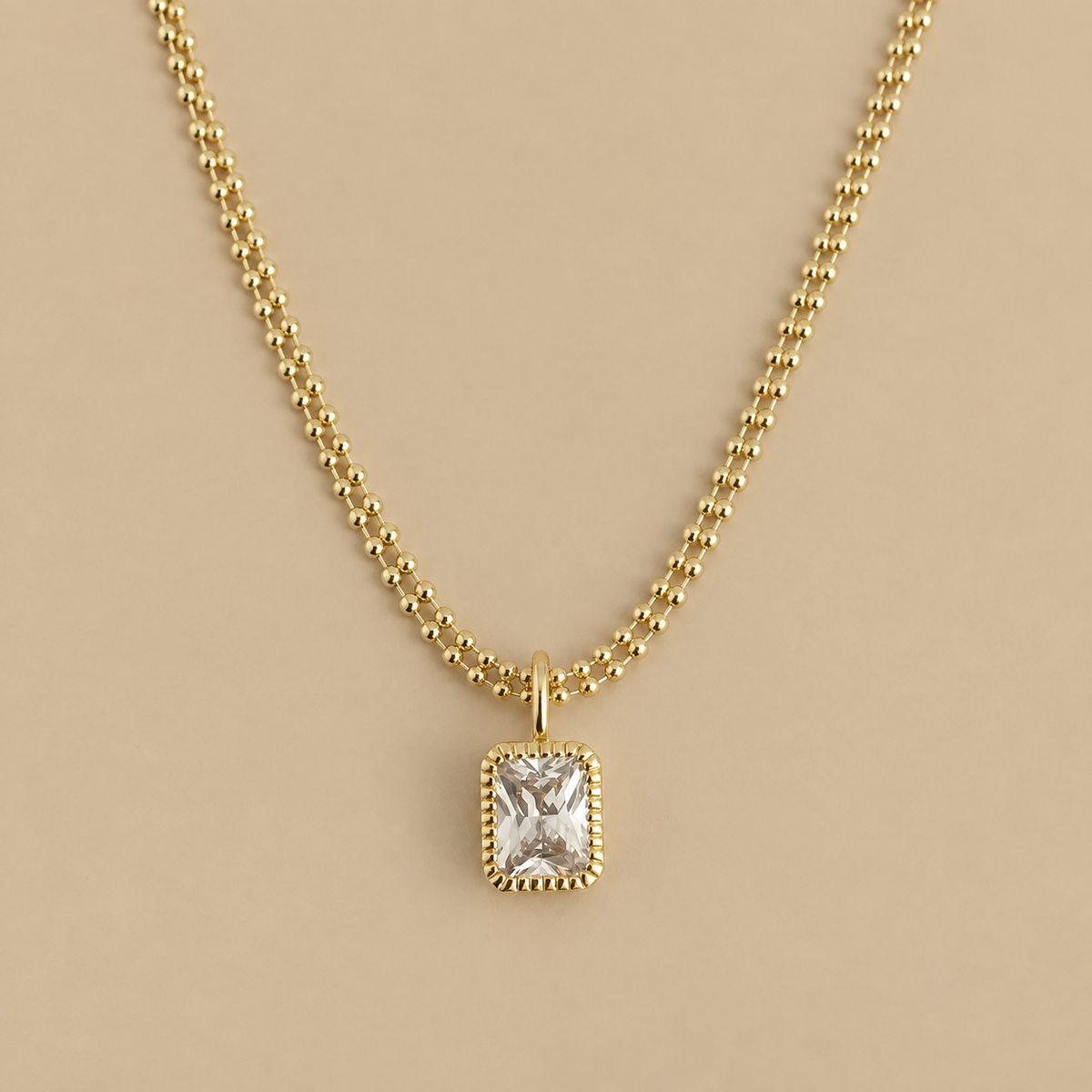 A gold chain necklace with a rectangular bezel-set clear gemstone pendant, displayed against a solid beige background.