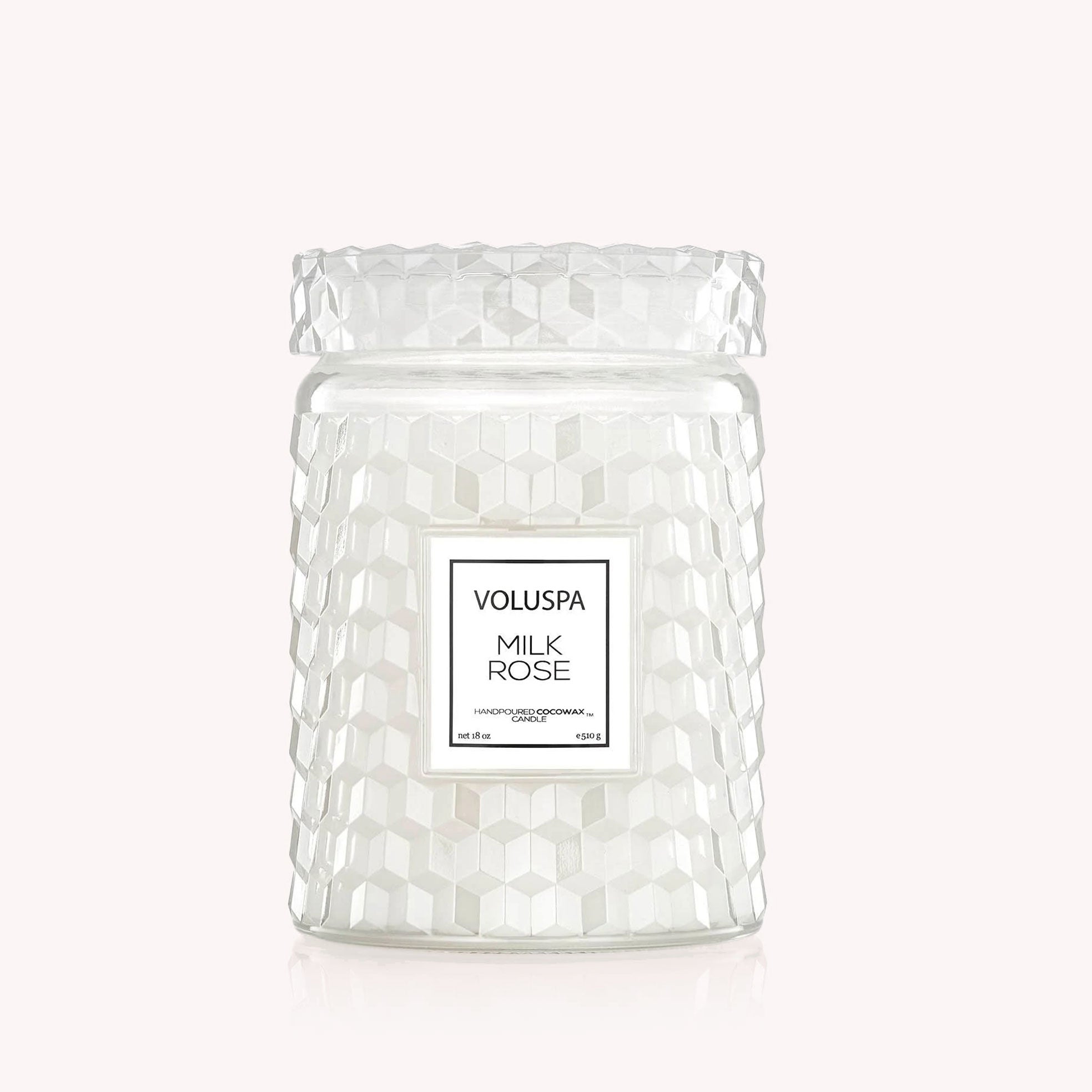On a light pink background is a white glass jar candle with a milky effect to the glass along with a lid and a rectangular label in the center that reads, "Voluspa Milk Rose".