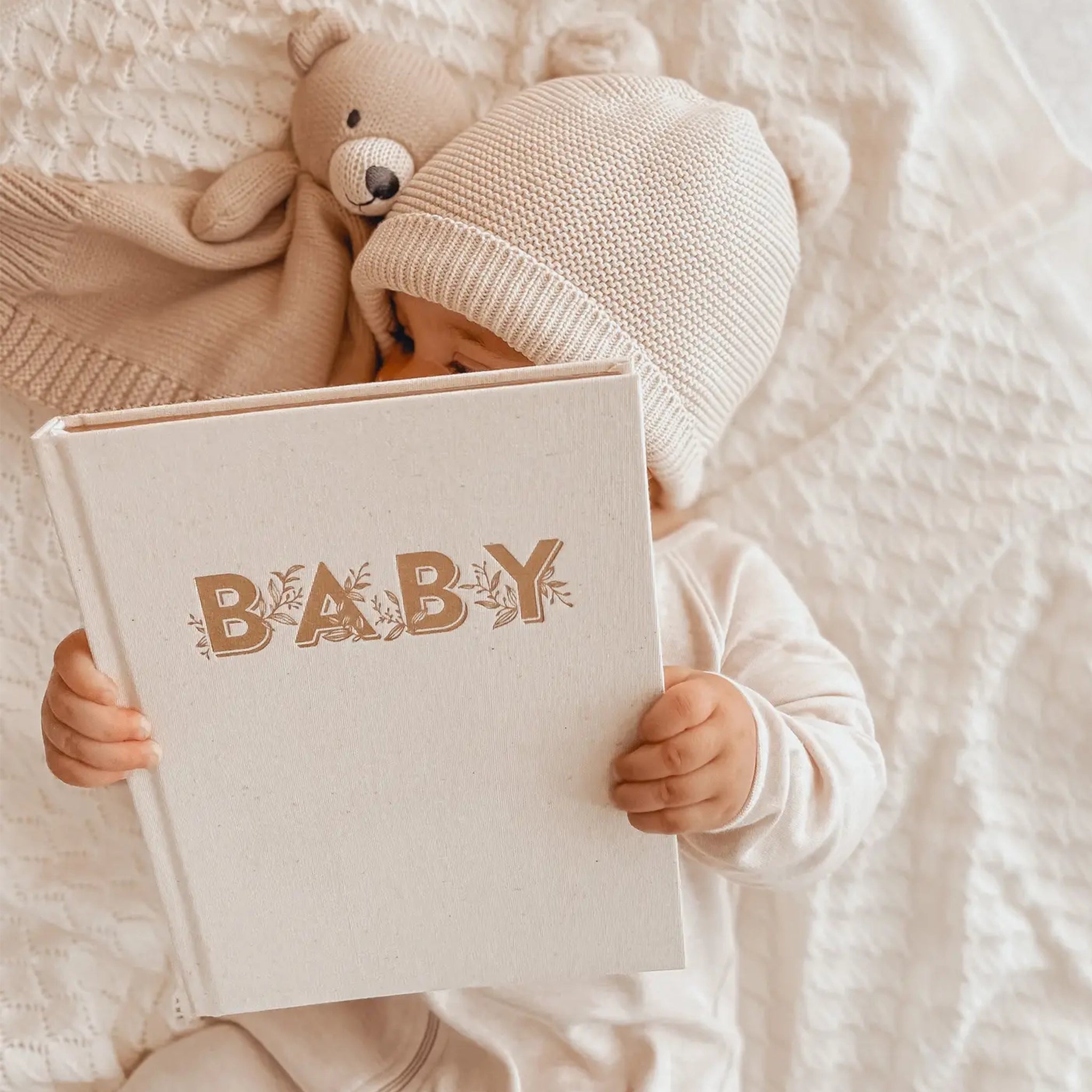 A model baby holding a neutral ivory colored book with gold lettering on the front that reads, &quot;Baby&quot;. 