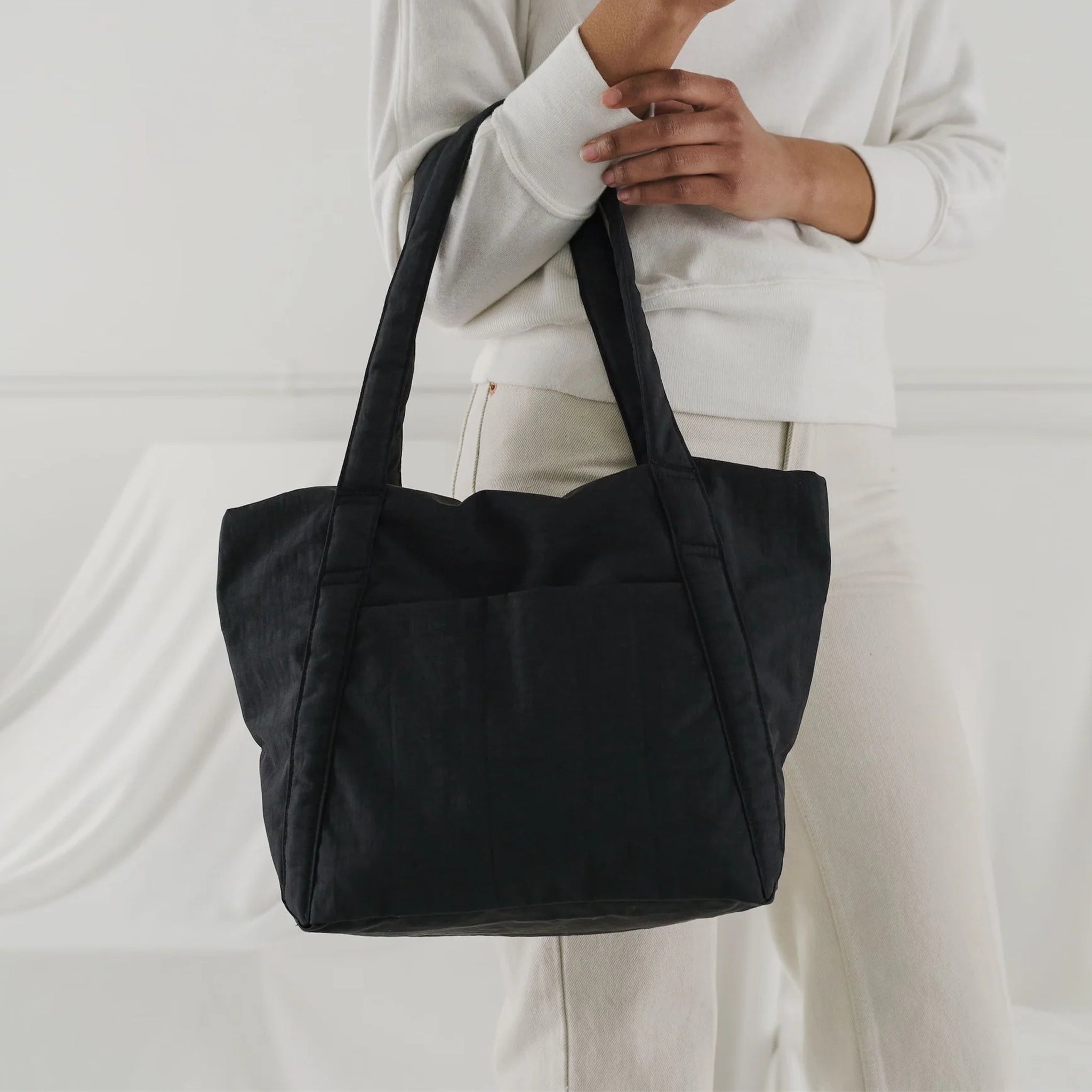 On a white background is a model holding the Mini Cloud Bag in the shade black. It is a rectangular shaped puffy bag with shoulder straps and a wide opening. 