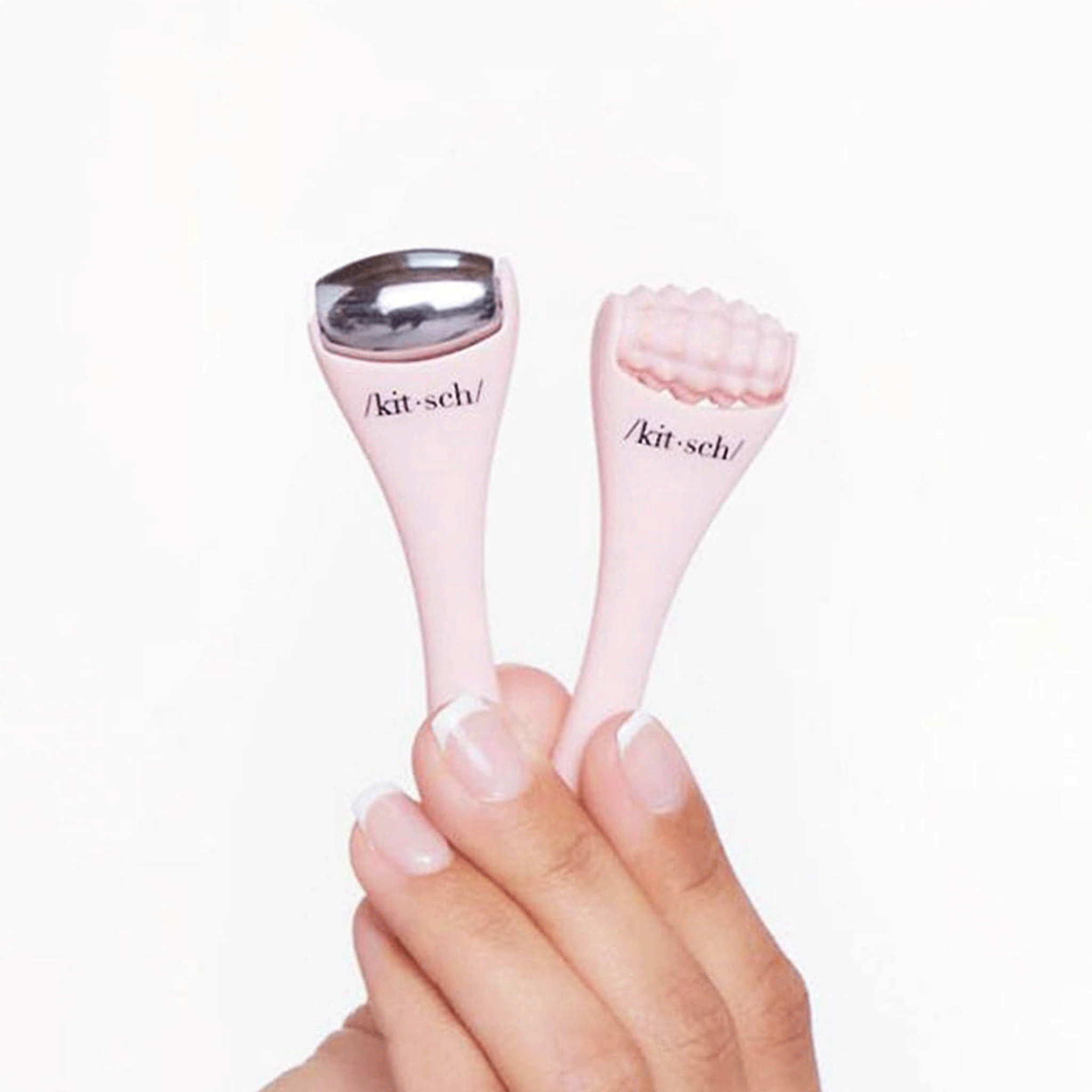 On a white background is a model&#39;s hand holding two mini facial massage tools. One with a smooth stainless steel roller and the other with a more textured light pink roller.