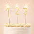 On a light pink background is a photo of three gold sparkling candles in the shape of a "1" a "2" and a "3".