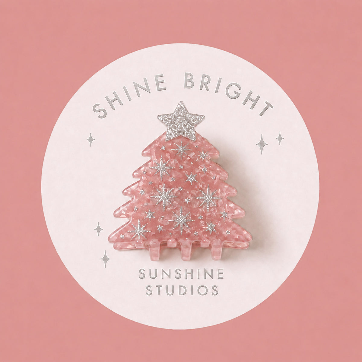 A pink Christmas tree-shaped mini hair clip with silver glittery snowflakes and a silver star topper, displayed on a white circular backing card that reads 'SHINE BRIGHT SUNSHINE STUDIOS' against a dusty pink background.