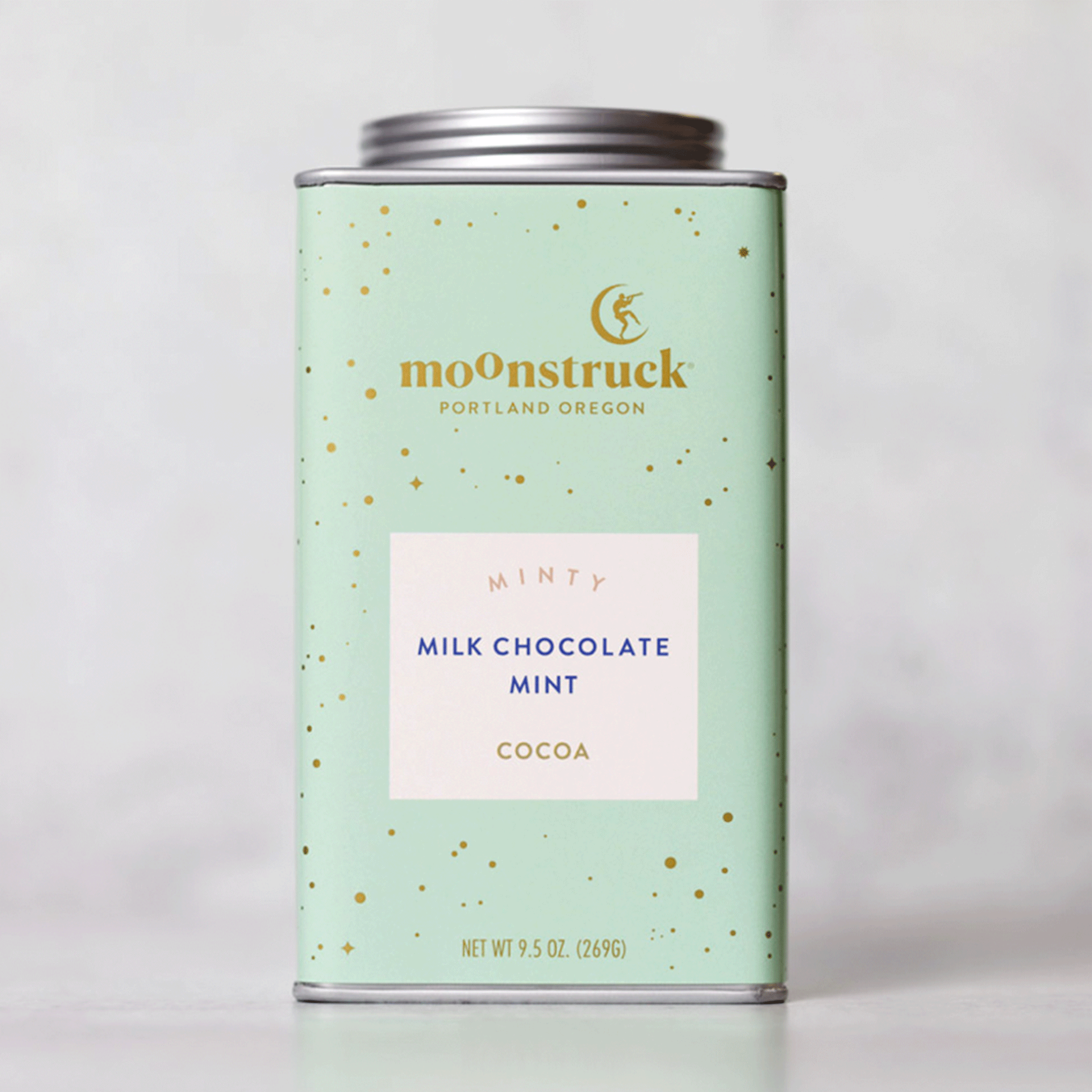 On a grey background is a mint green tin with gold sparkle details and text that reads, &quot;moonstruck Portland Oregon&quot;, &quot;Minty Milk Chocolate Mint Cocoa&quot;.