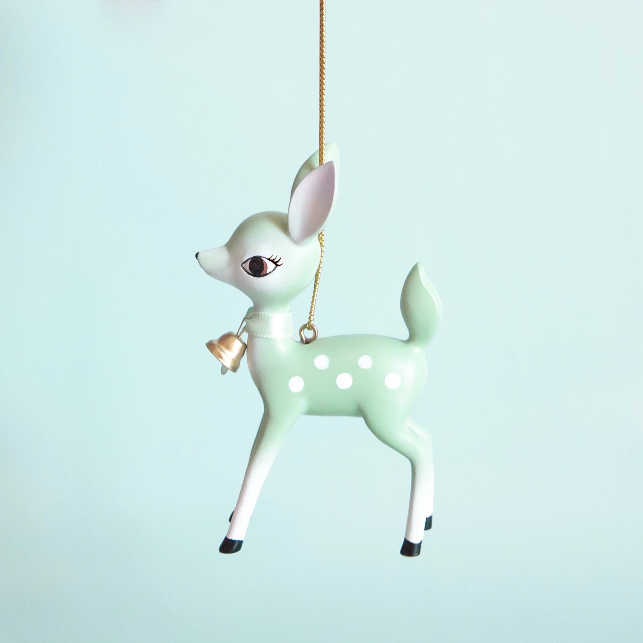 On a light blue background is a mint green retro deer ornament with white details and spots and a gold bell on the front of its neck along with a gold string loop for hanging.