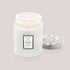 On a white background is a white decorative glass jar with a coordinating lid and a label on the front that reads, "Voluspa Mokara"