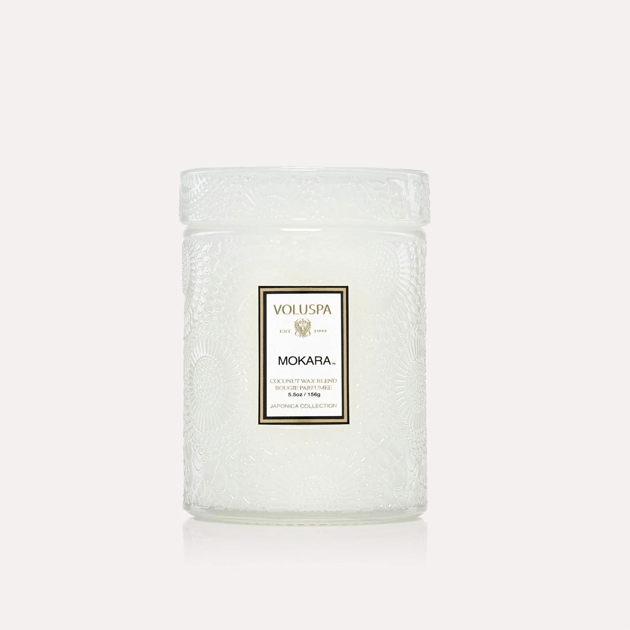 A decorative glass jar candle in a white color with a rectangular label on the front that reads, "Voluspa Mokara" with a coordinating decorative glass lid.