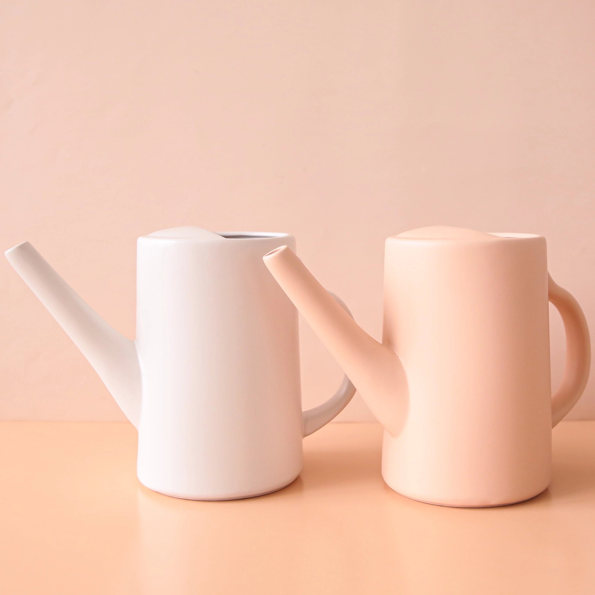 On a peachy background is two ceramic watering cans in a blush color and white. They have narrow spouts and curved handles. 