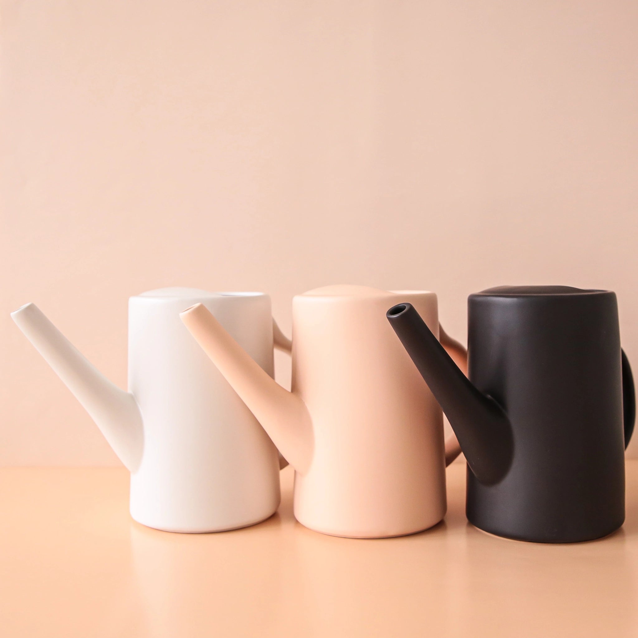 On a peachy background is three ceramic watering cans in a blush color as well as white and black. They have narrow spouts and curved handles.
