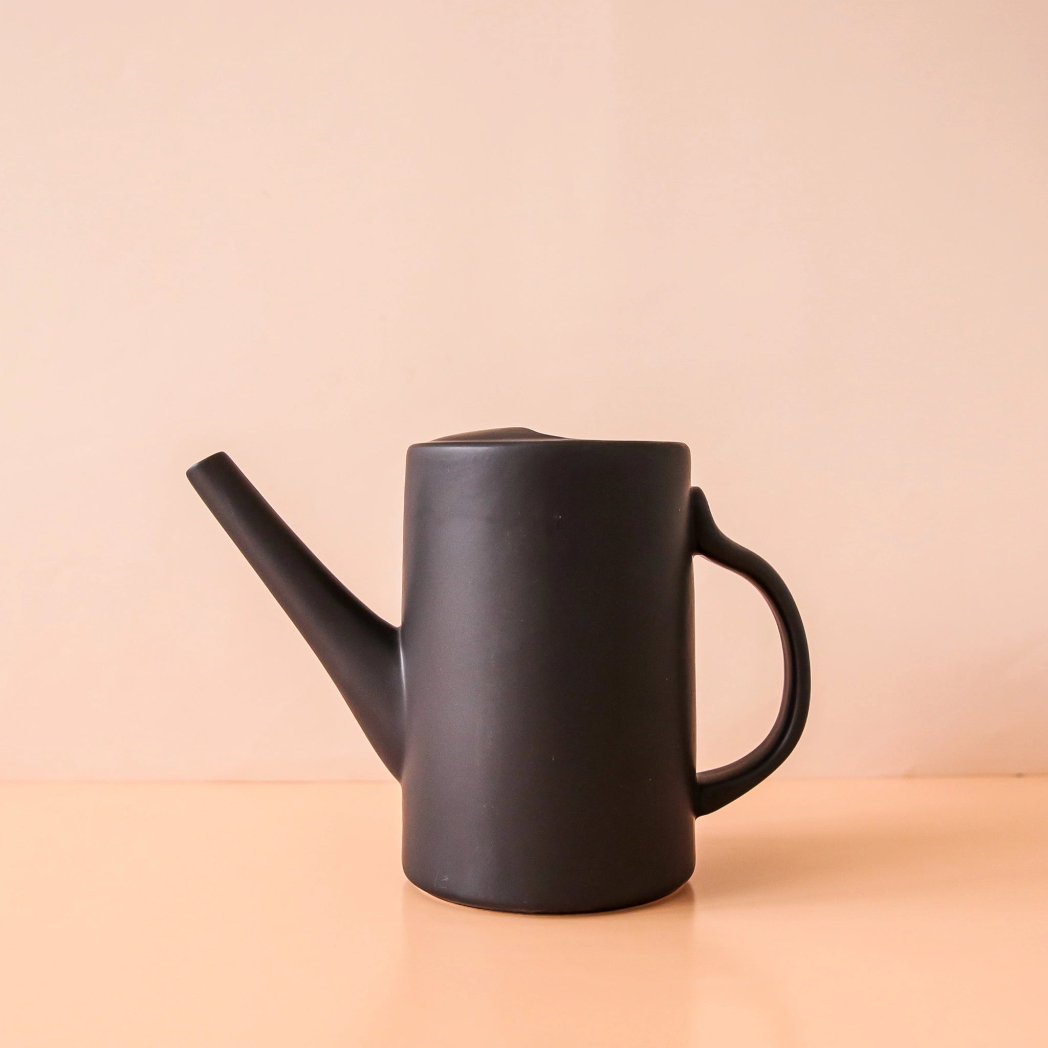 On a peach background is a back ceramic watering can with a narrow spout and a curved handle.