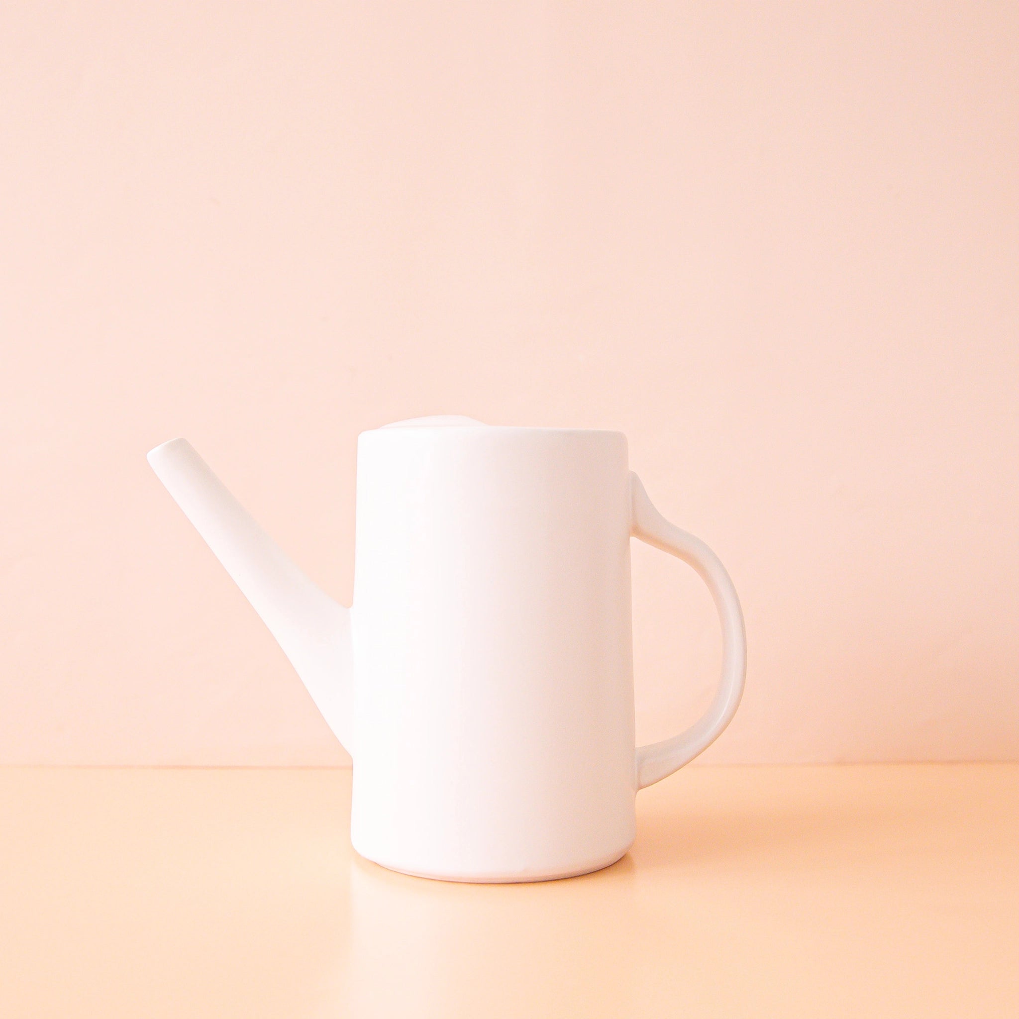 On a peach background is a white ceramic watering can with a narrow spout and a curved handle.