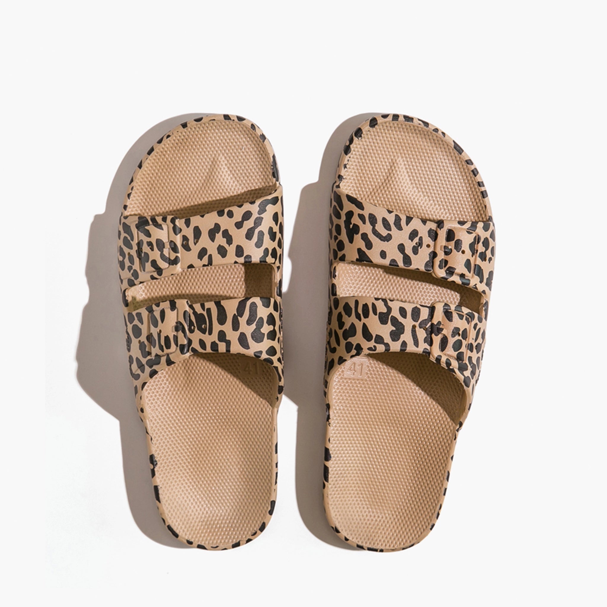 On a white background is a pair of leopard print sandals with two straps across the front. 