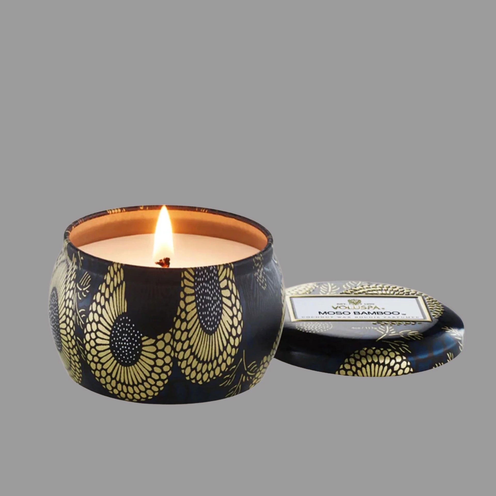 On a gray background is a black and gold decorative tin single wick candle with a label on the lid that reads, &quot;Voluspa Moso Bamboo&quot;.