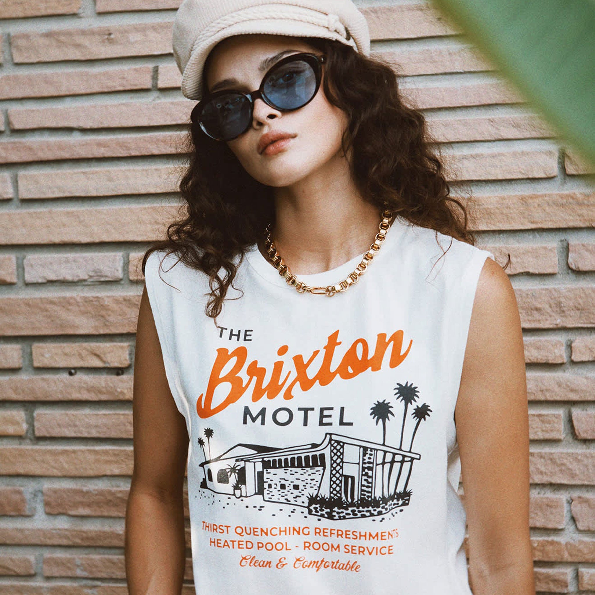 Model is wearing a white tank top, and is styled with a beige hat and black sunglasses. 