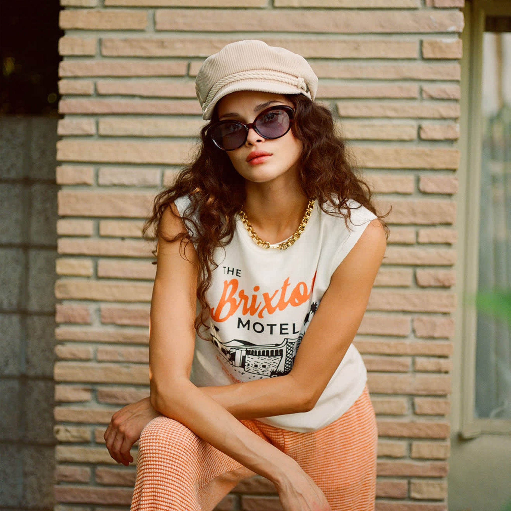 Model is wearing a white tank top, and is styled with a beige hat and black sunglasses. 