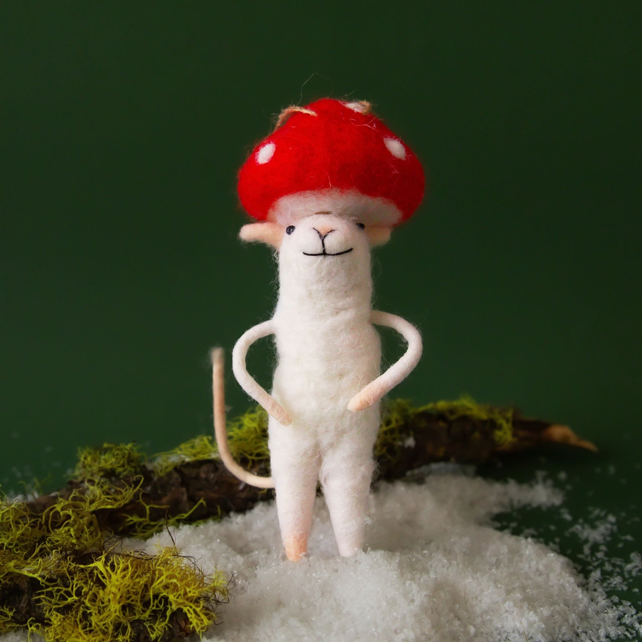 On a dark green background is a white felt mouse ornament with a red mushroom hat on and staged on a snowy and mossy backdrop. 