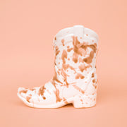 On a peachy background is a brown and white ceramic cowboy boot shaped candle. 