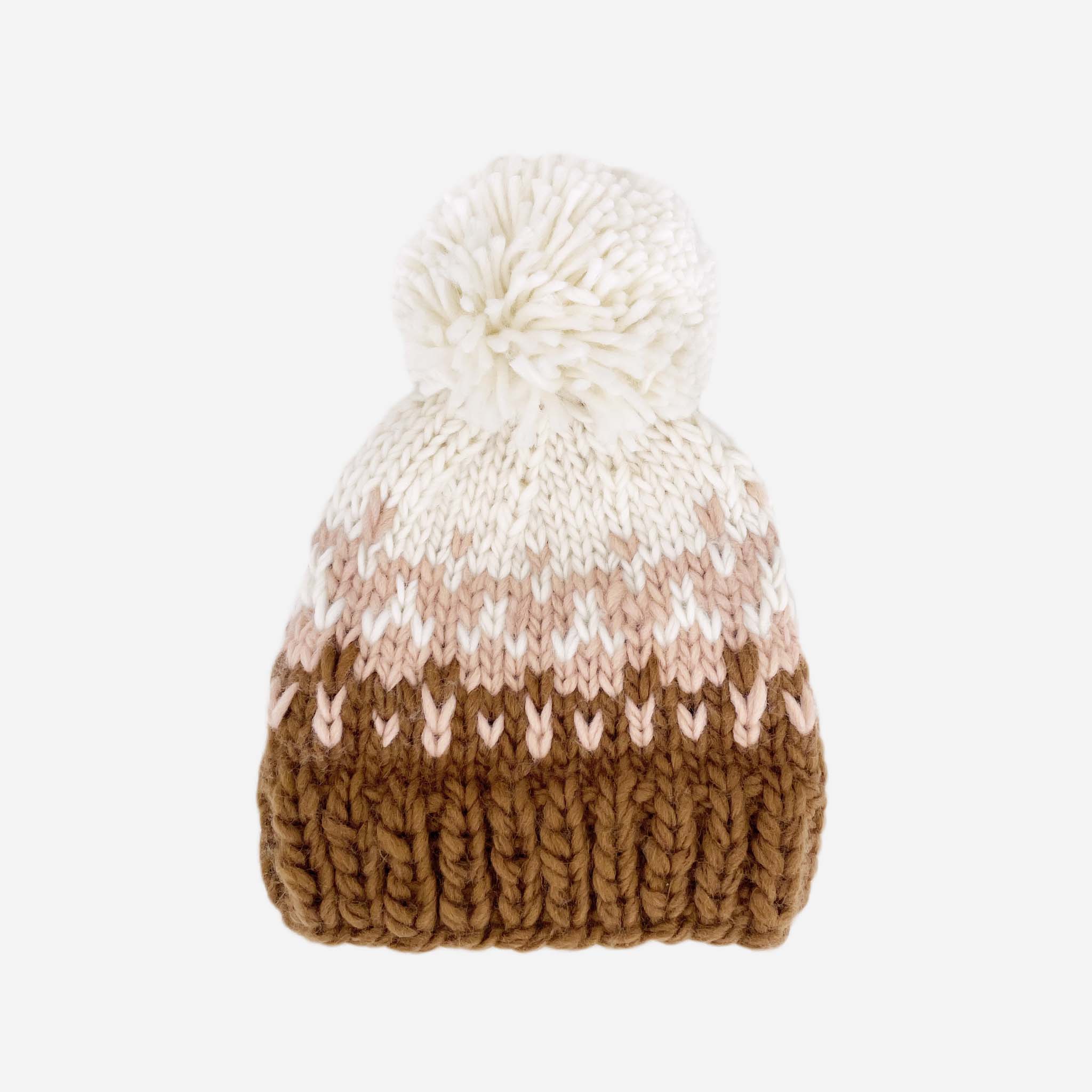 On a white background is a children&#39;s knit beanie with brown, light pink and ivory colors along with an ivory pom on the top.
