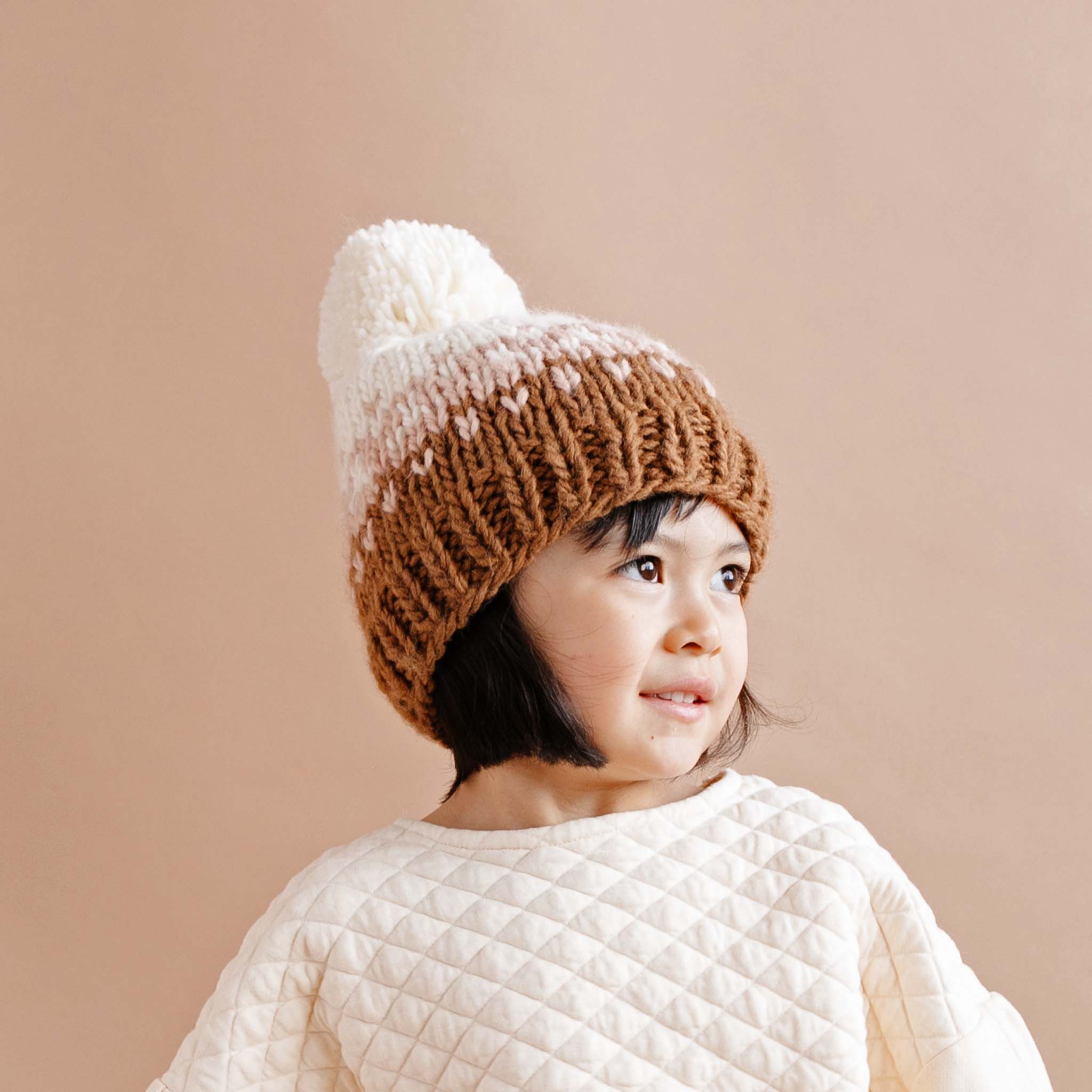 On a tan background is a children&#39;s knit beanie with brown, light pink and ivory colors along with an ivory pom on the top.