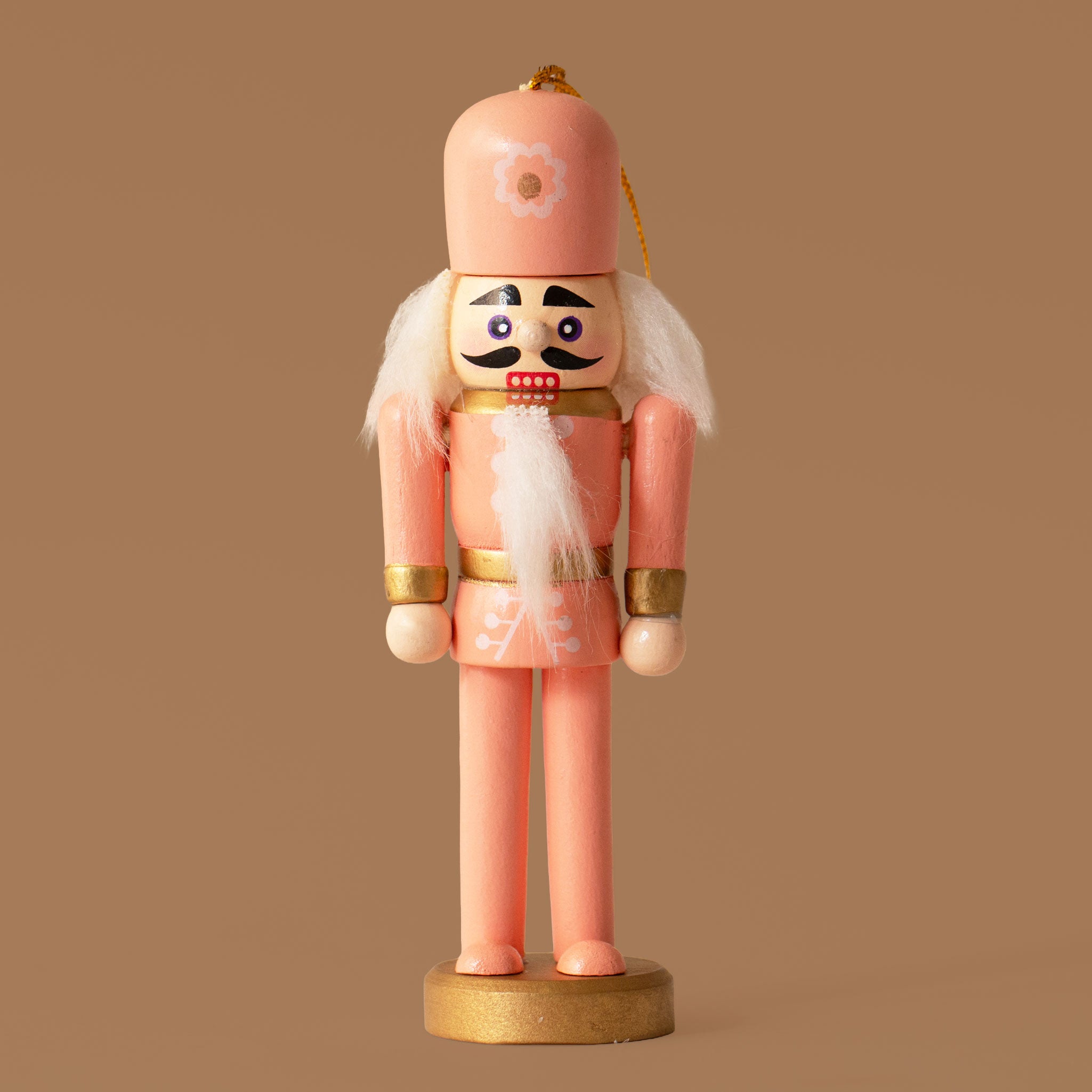 A coral colored wooden nutcracker figure with a tall pink hat, white hair and beard, gold accents, and a gold round base, standing against a warm brown background.