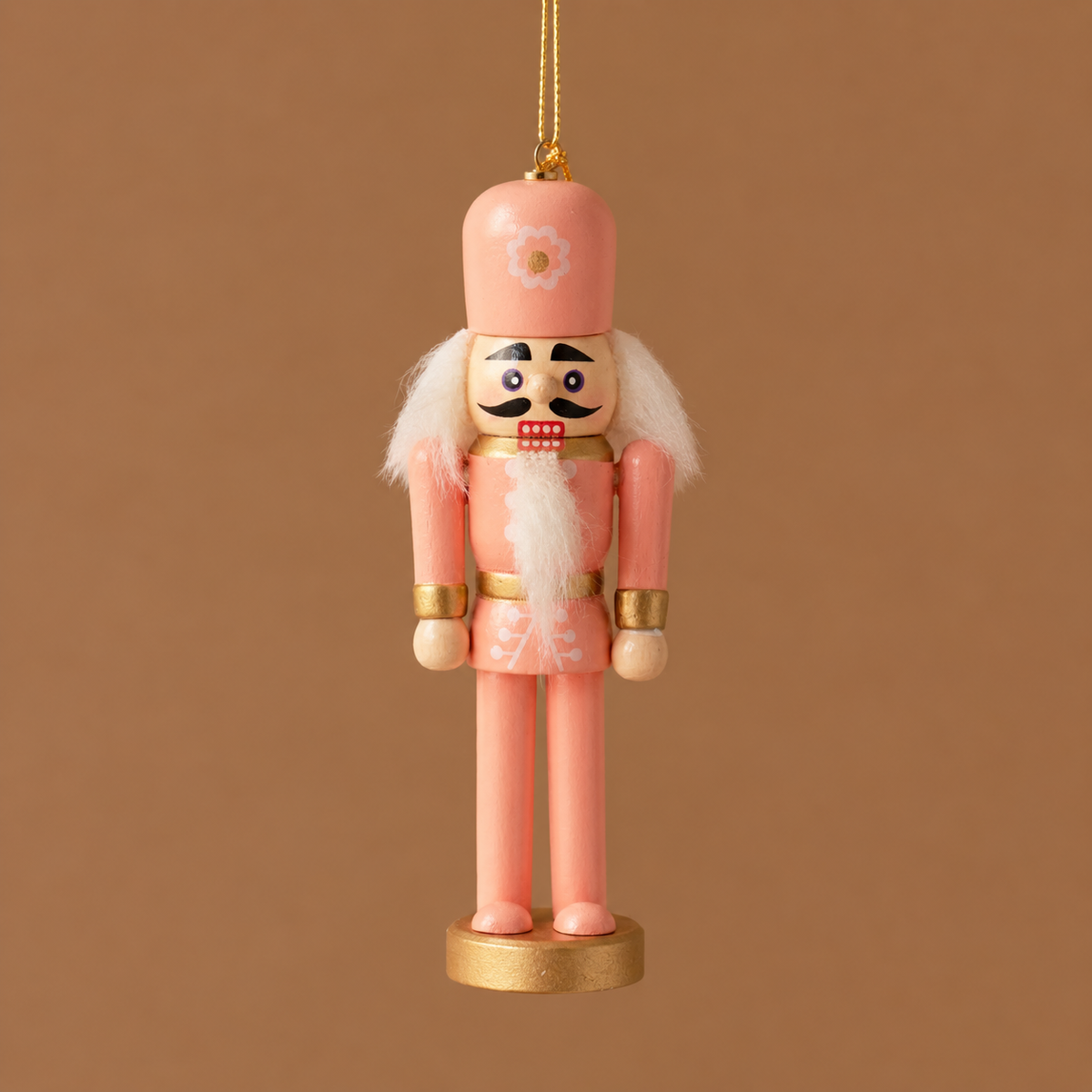 A pink wooden nutcracker ornament hanging against a solid warm brown background. The ornament features a pink uniform, white faux fur details, a gold-plated base and belt, and a painted face with a black mustache.