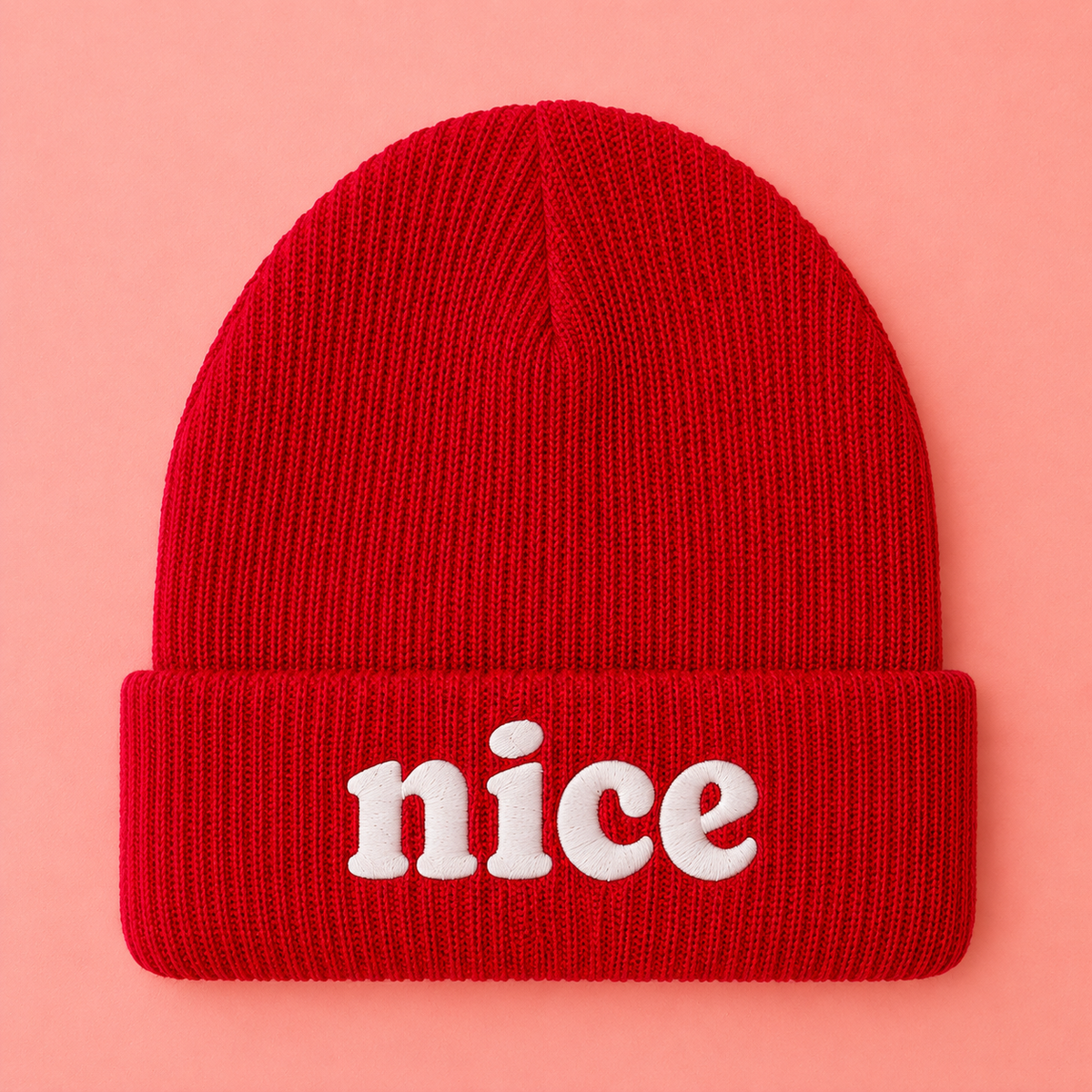 A bright red ribbed knit beanie with the word 'nice' embroidered in white on the front cuff, shown against a solid pink background.