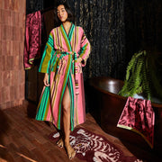 Person wearing a striped robe in pink, green, and olive standing in a bathroom with a copper tub and patterned rug.
