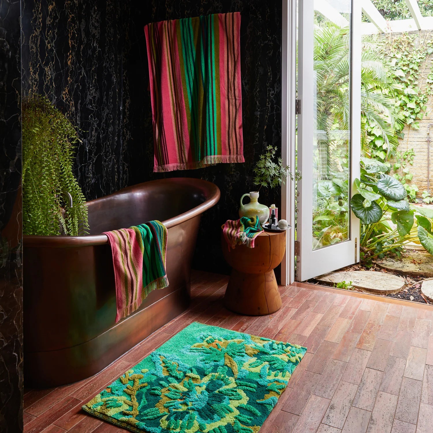 Freestanding copper bathtub in a dark marble bathroom with colorful striped towels, a green patterned bath mat, and an open door leading to a lush garden.