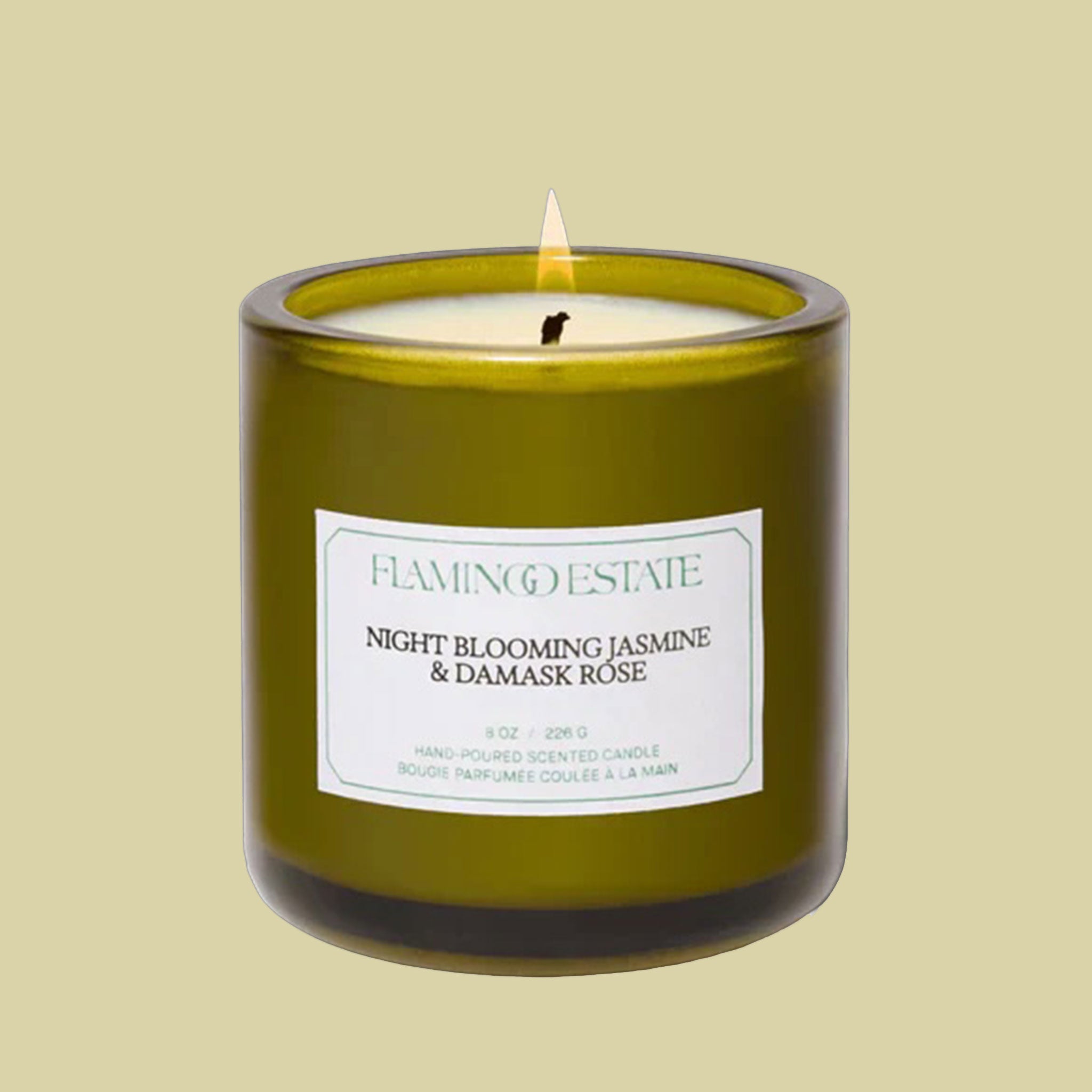 A green glass candle with a white label that reads, "Night Blooming Jasmine & Damask Rose"