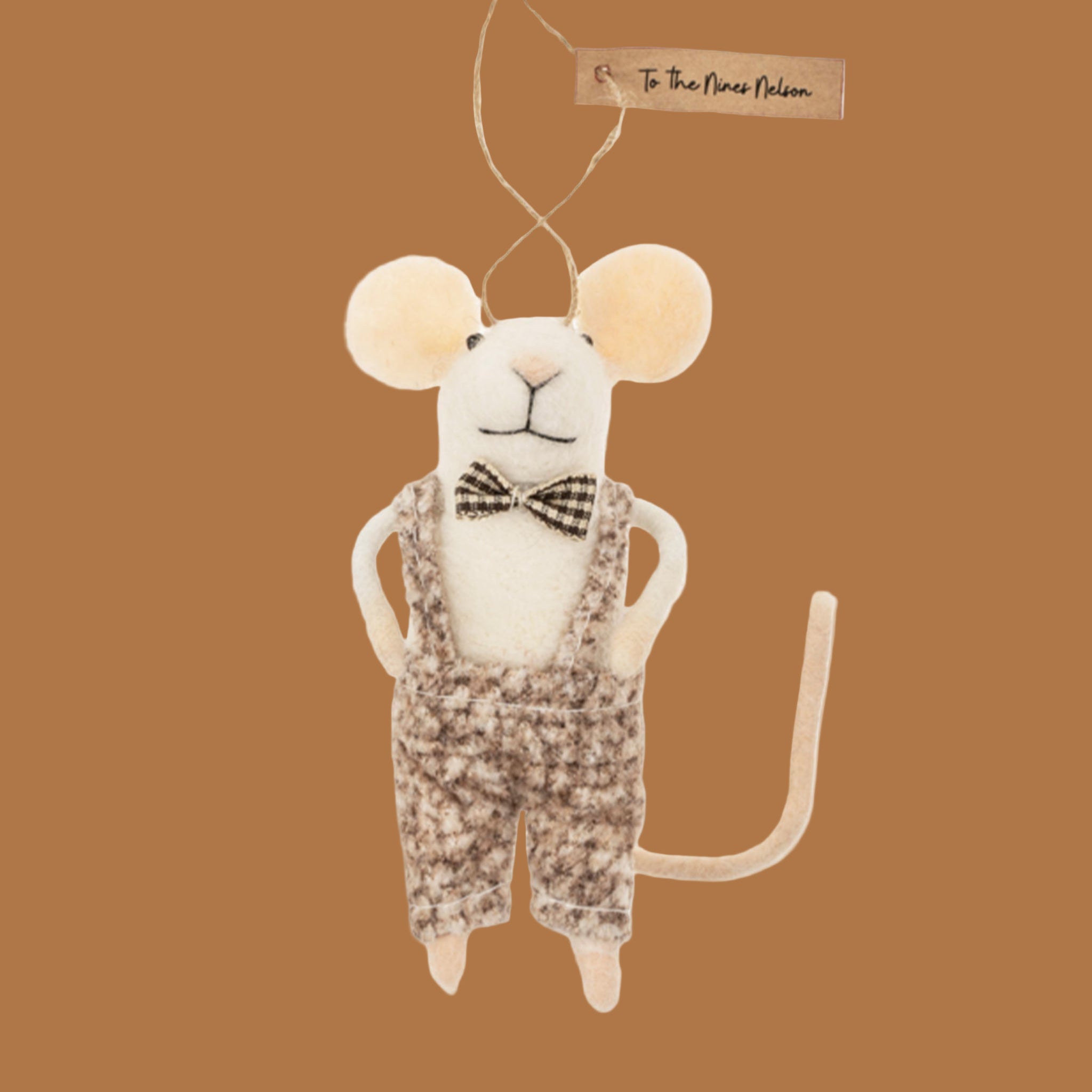 A felted mouse ornament wearing a black and white bow tie with overalls stands on a tan backtround.