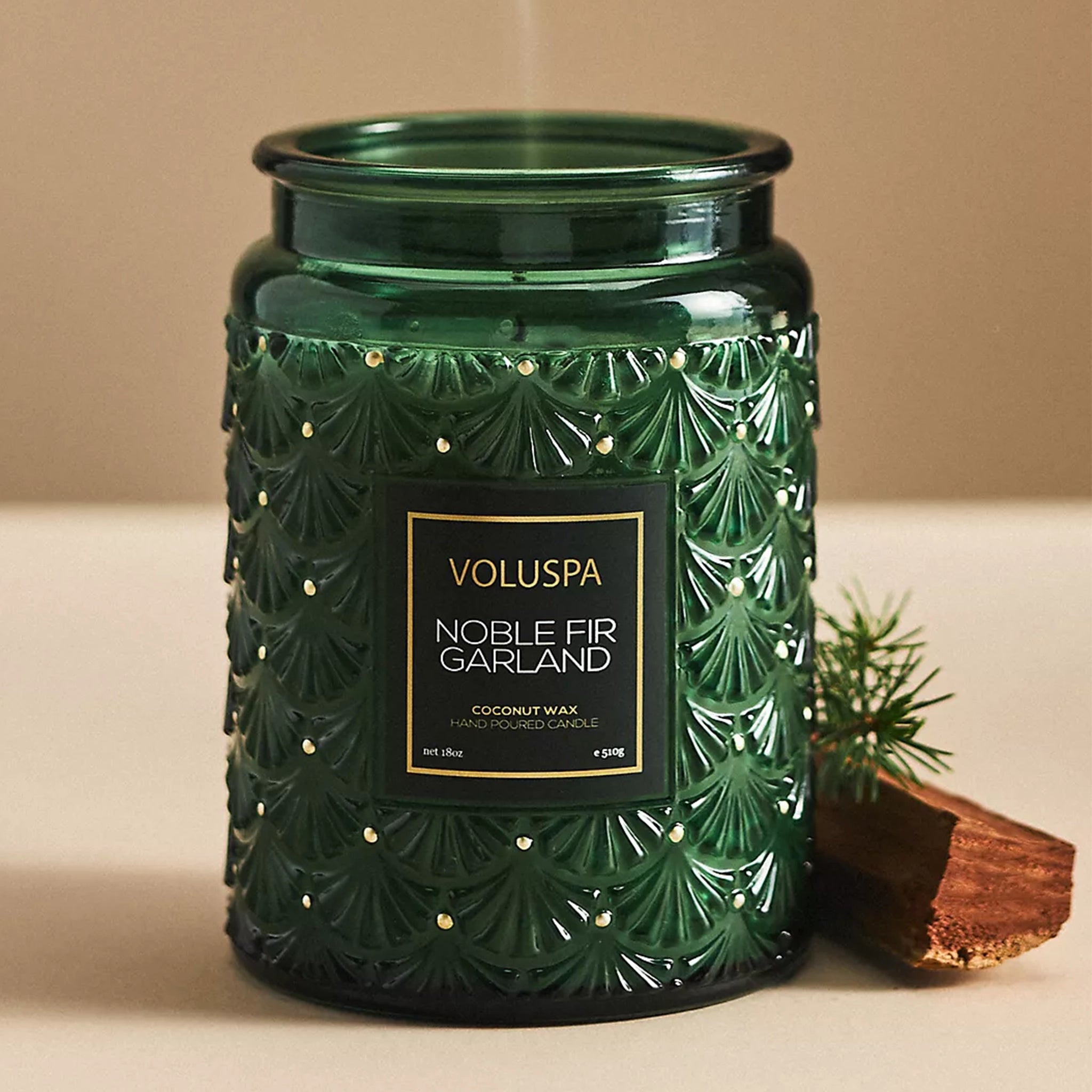 On a tan background is a dark green decorative glass jar candle with gold dot details and a label in the center that reads, "Noble Fir Garland".