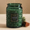 On a tan background is a dark green decorative glass jar candle with gold dot details and a label in the center that reads, "Noble Fir Garland".
