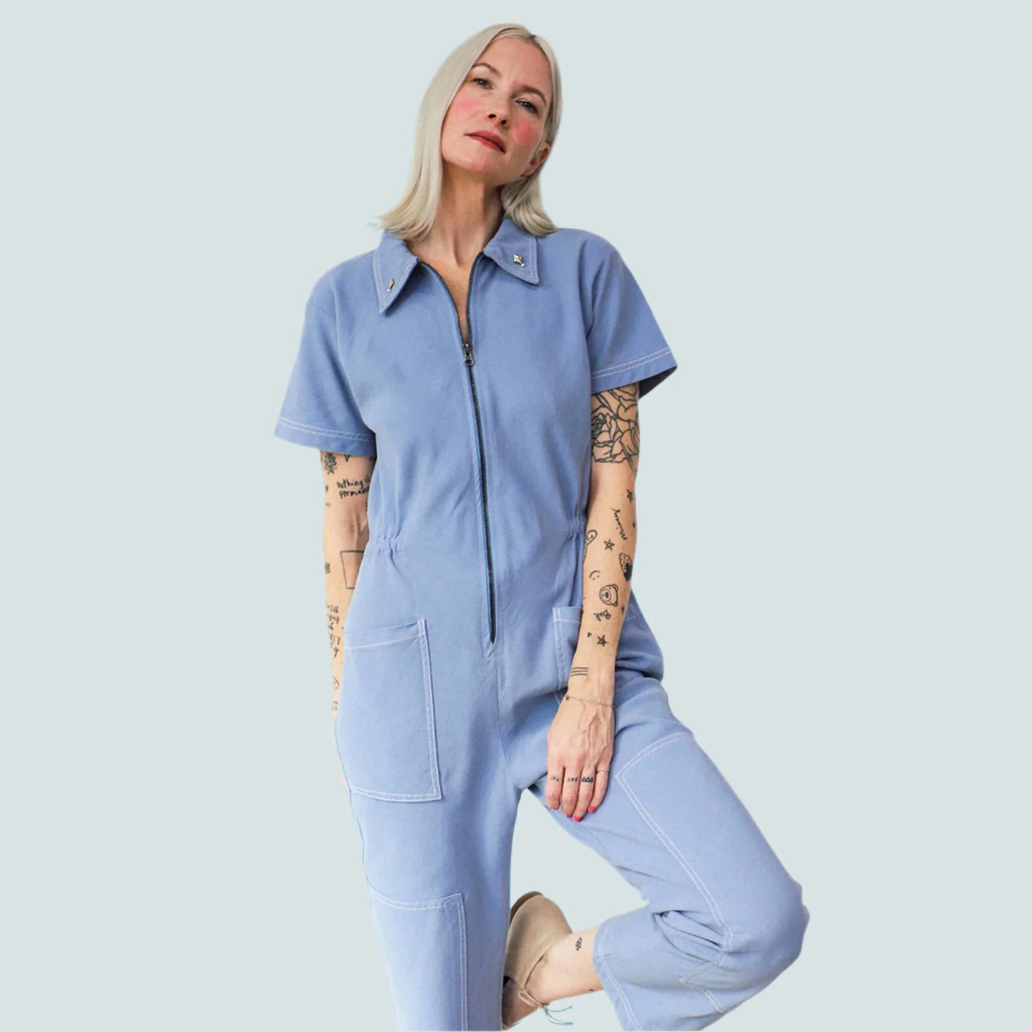 A woman with blonde hair wears a blue jumpsuit on a light blue background.