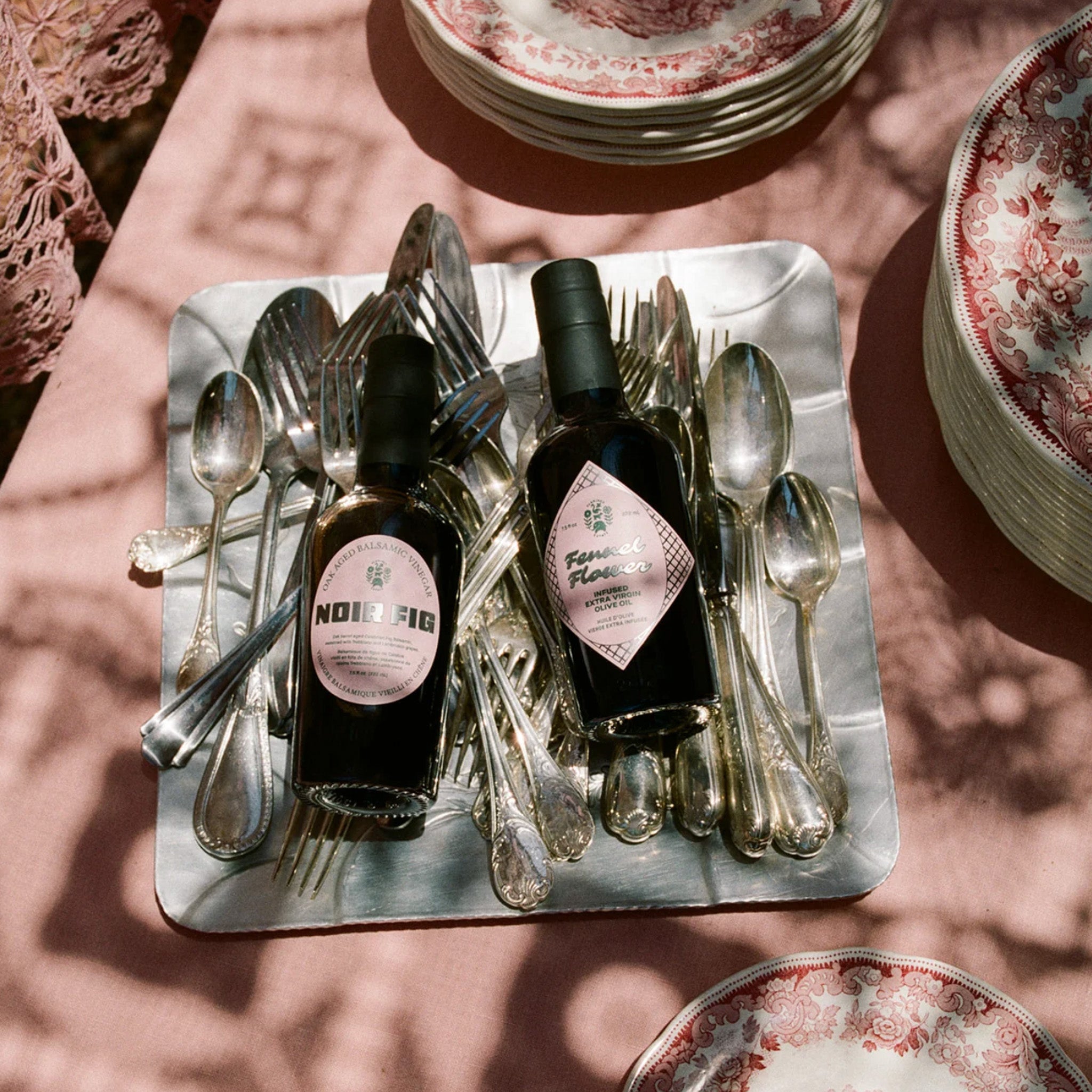 Two bottles of balsamic oil on a table filled with silverware, a pink tablecloth and ceramic dishes.