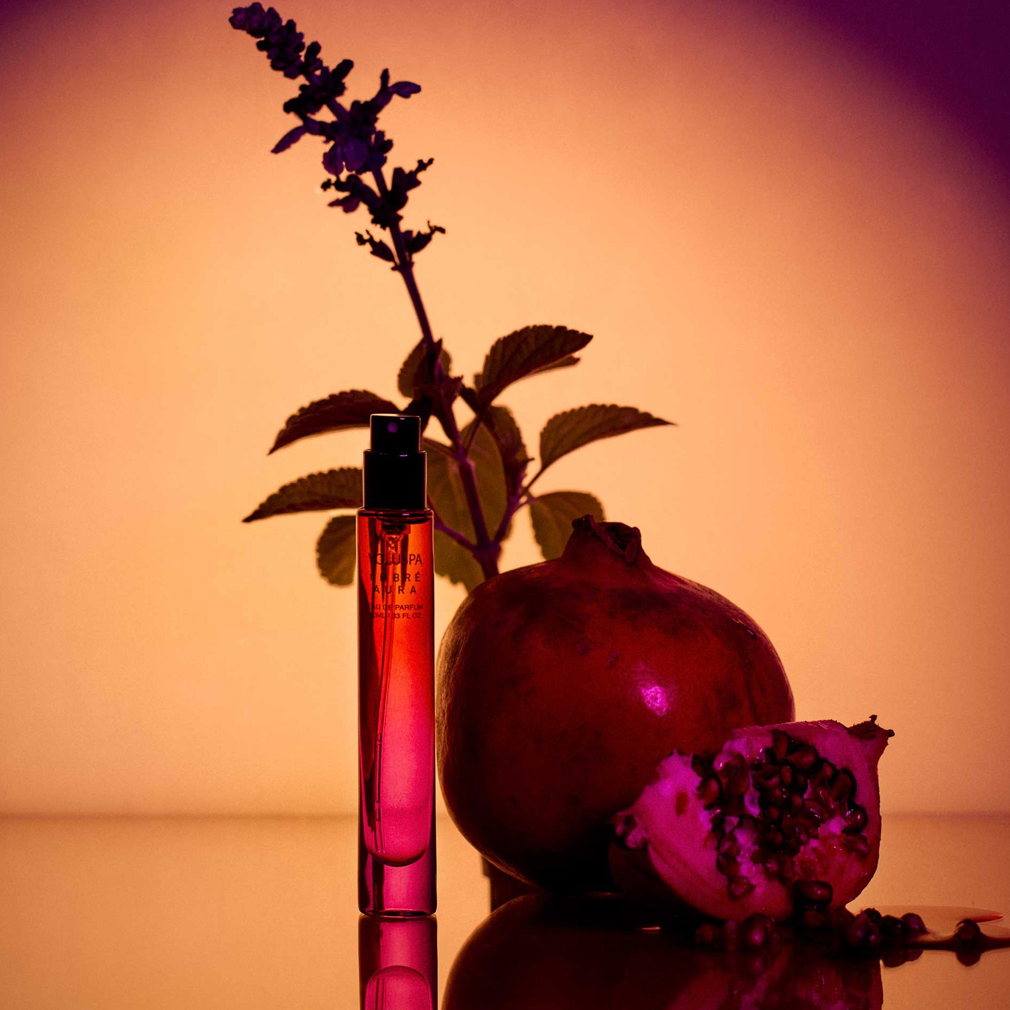 A dark purple, red ombre travel bottle of perfume with black details and a black label with gold text that reads, 'Voluspa Ombre Aura'. 