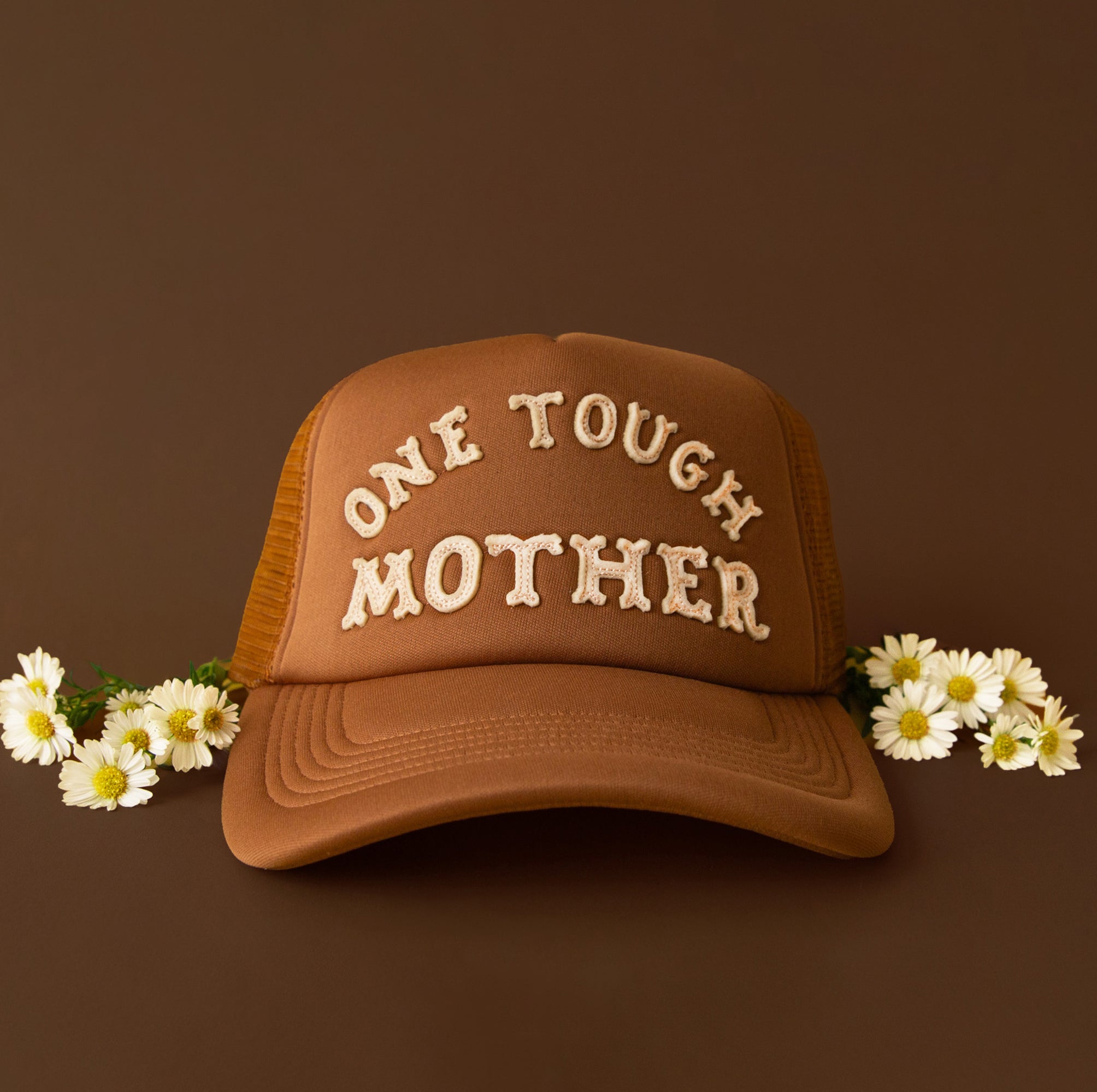 Brown trucker hat with embroidered text reading “One Tough Mother,” styled with small white daisies on either side against a warm brown background.