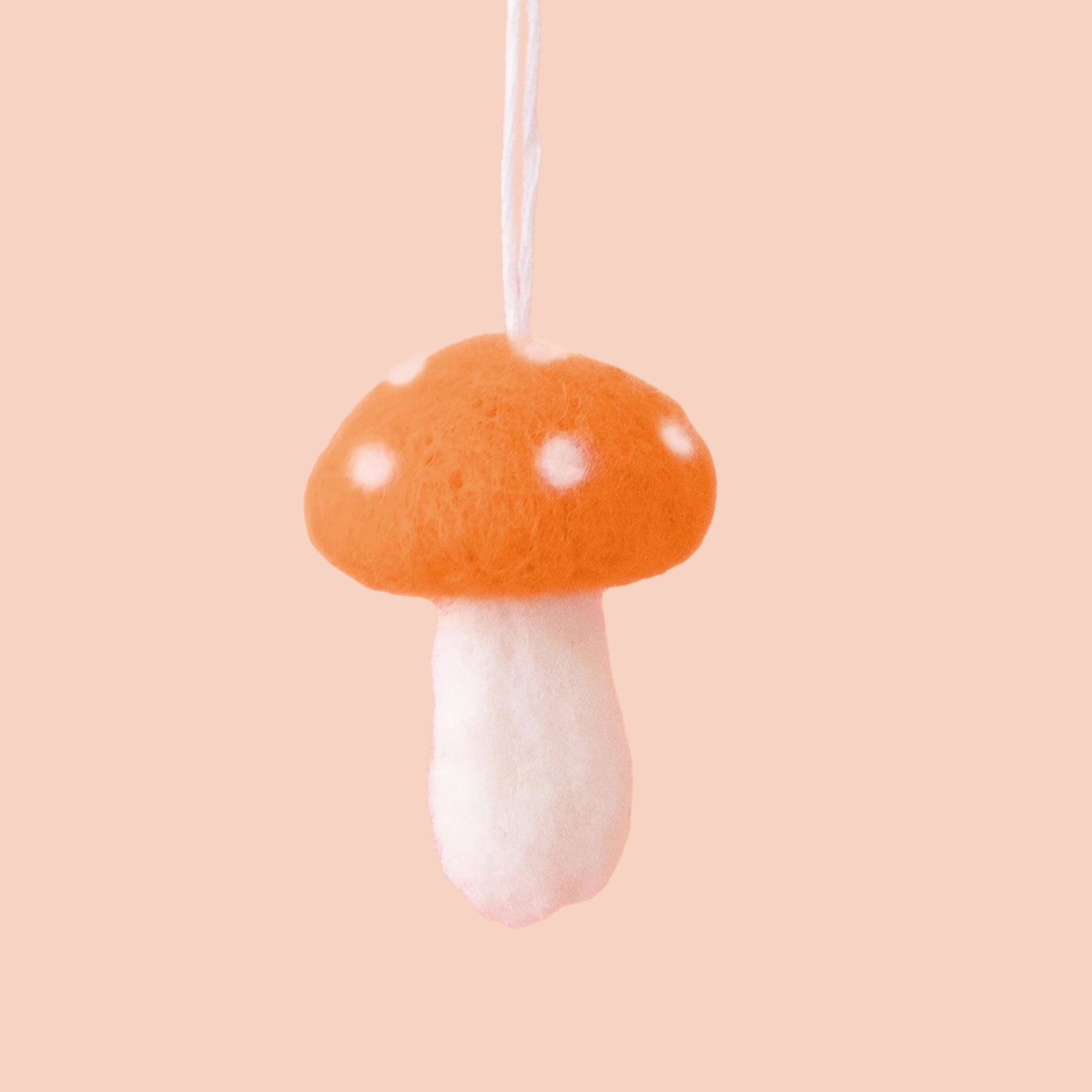 Orange felt mushroom ornament with white spots on the mushroom cap, and a white stem are shown against a peach colored background. 