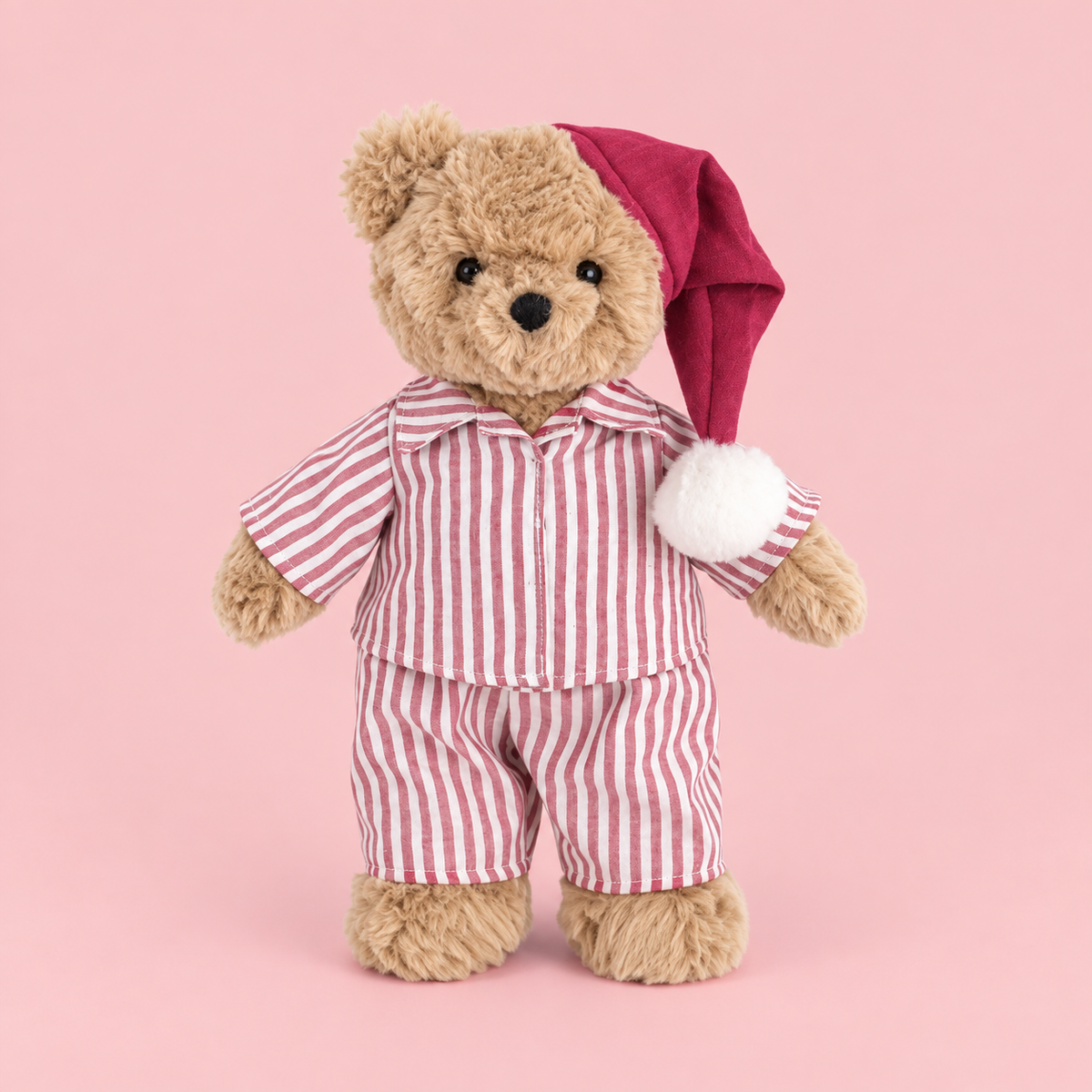 A plush teddy bear wearing red and white vertically striped pajamas and a dark red Santa hat with a white pom-pom, standing against a solid pink background.