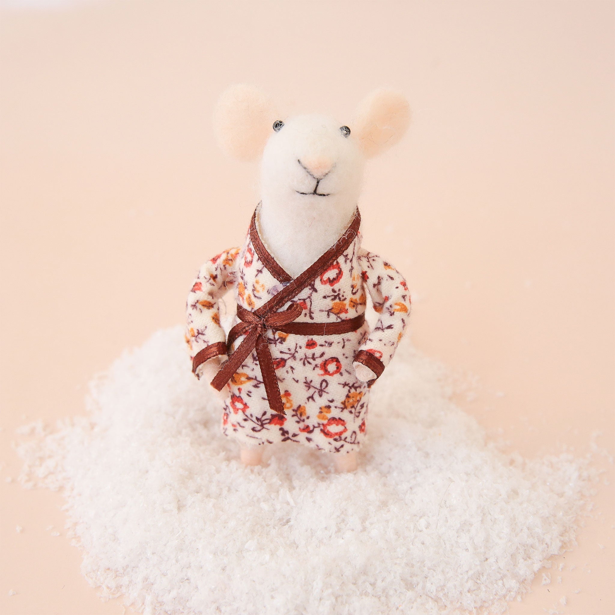 On a peach background is a white felt mouse ornament with a floral pyjama robe on. 
