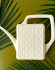 An ivory plastic watering can with a narrow spout and square handle and a rectangle breeze block design on the sides. The watering can is photographed on a lime green ground with palm fronds spilling into the image.