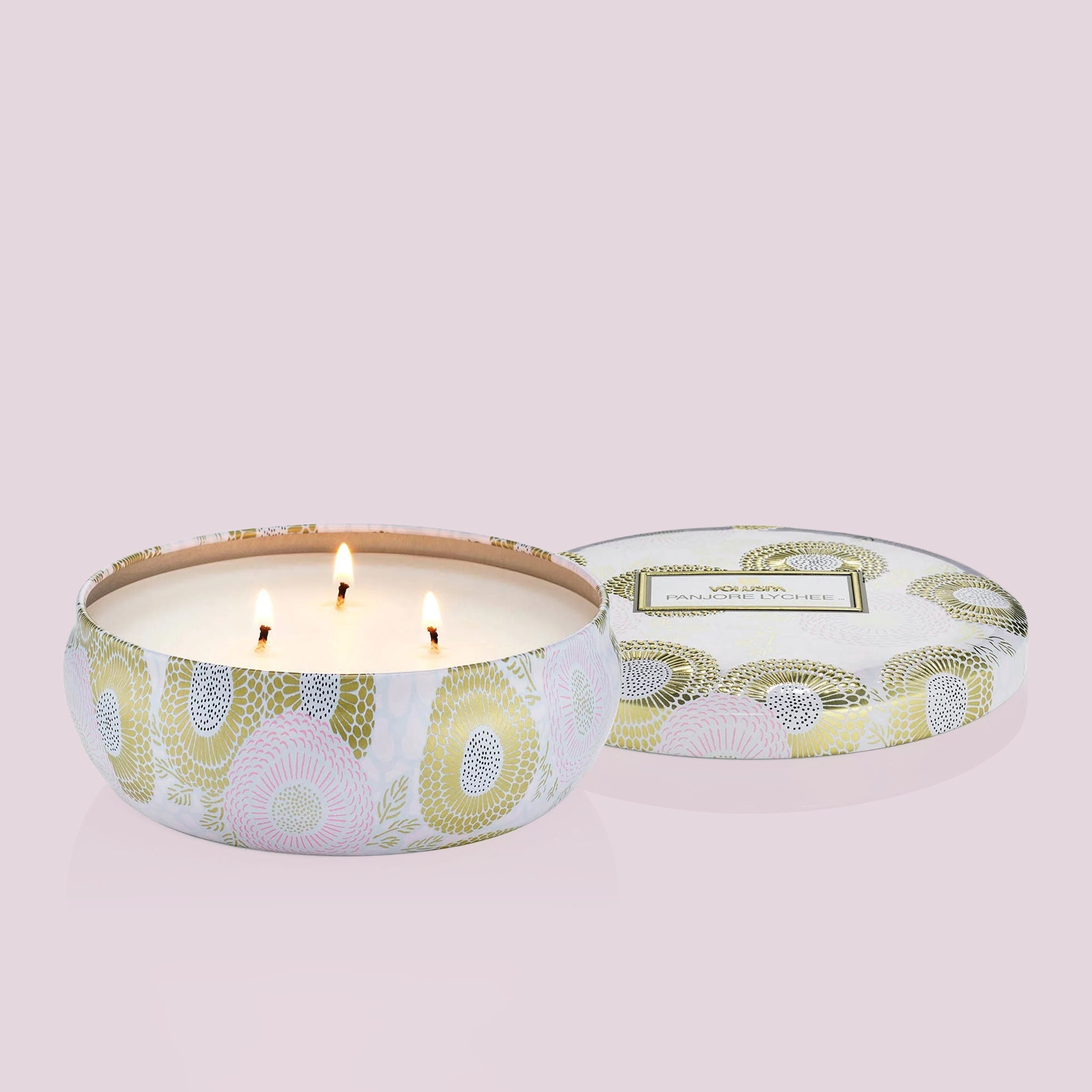 Inside the tin is a white candle with three white wicks. Leaning against the right side is a round tin lid. The lid has a light pink and gold floral pattern on it. In the middle is a white sticker with gold and black text inside.