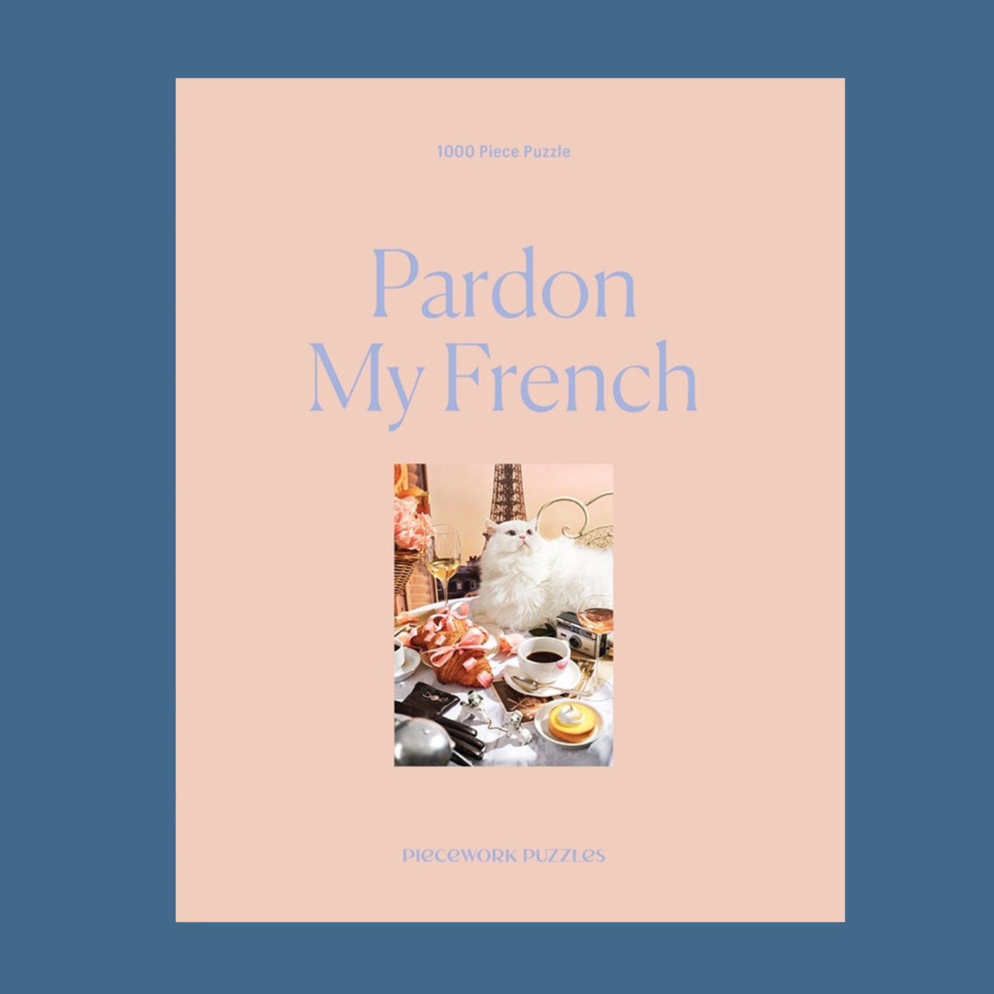 A peachy tan colored puzzle box with a 1000 piece puzzle inside with an image of a white fluffy cat surrounded by French goods such as croissants, macarons, champagne and the Eiffel Tower.
